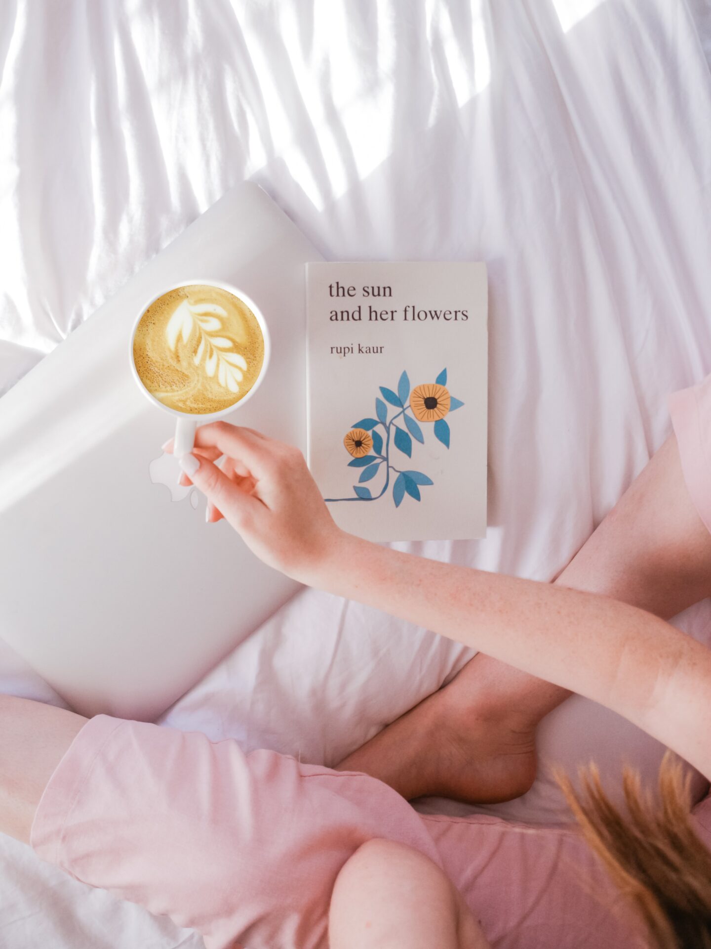 girl sat on a bed with a laptop, a book and mug of coffee