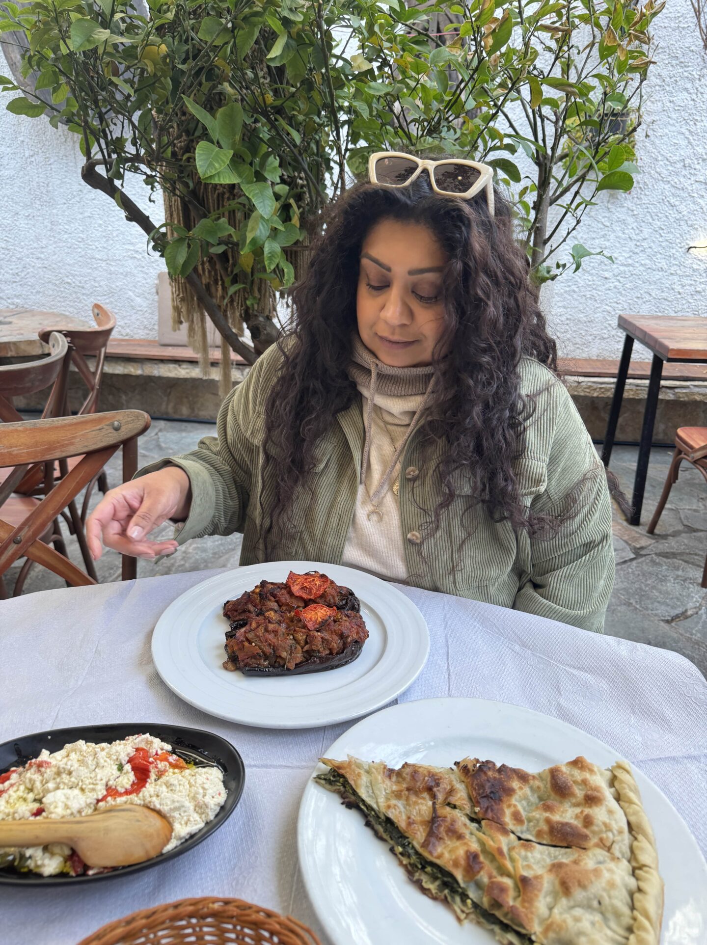 Nishi sitting outdoors at a table covered with traditional Albanian dishes, including lakror (a savoury pastry with spinach), a plate of stuffed aubergine topped with tomatoes, a dish of feta cheese with cherry tomatoes, and a basket of sliced bread. She is wearing a green jacket, cream hoodie, and white sunglasses resting on her head, with a leafy tree and rustic chairs in the background. The setting has a casual, garden-like ambiance