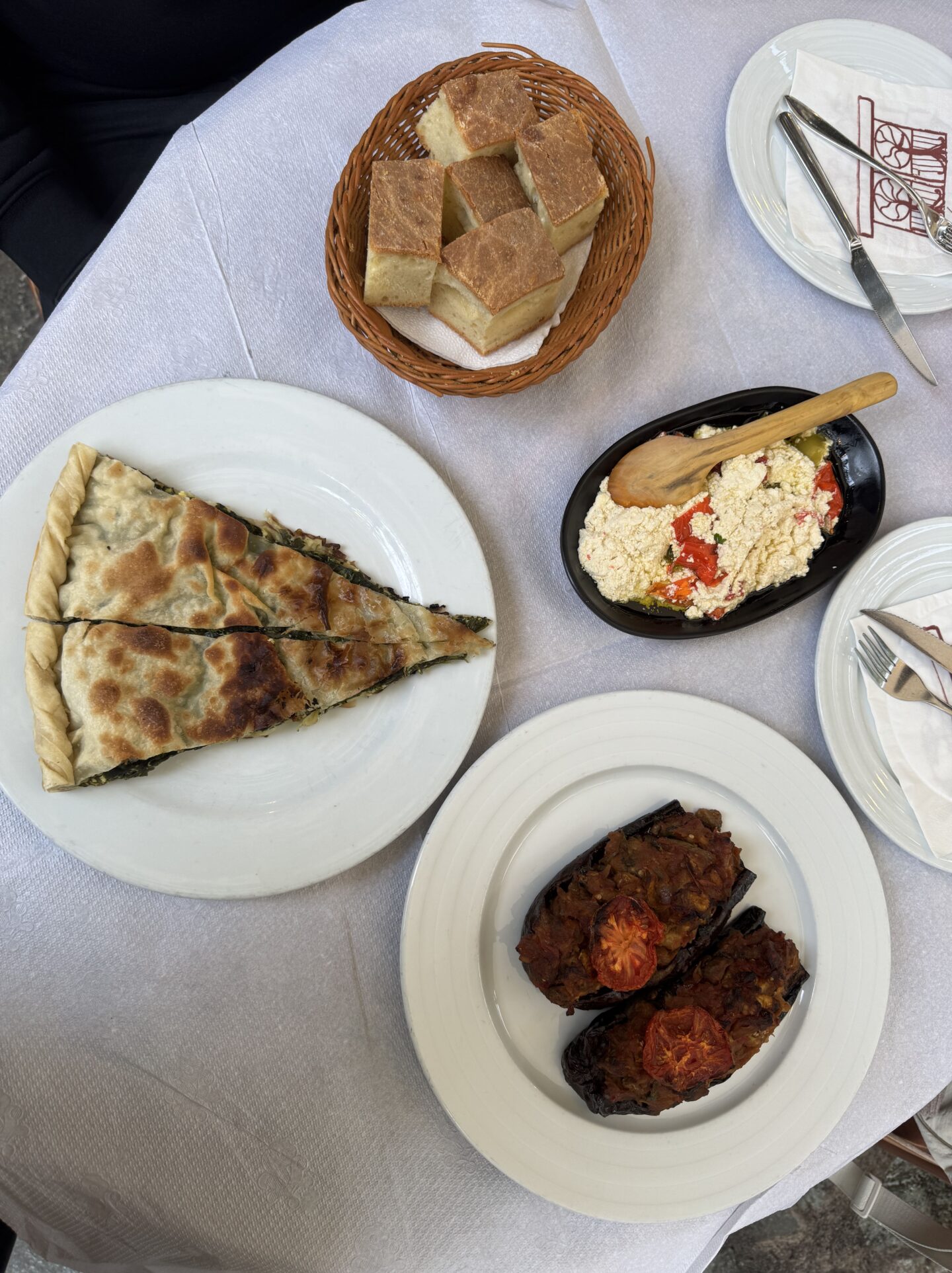 Top-down view of a table with traditional Albanian dishes. The spread includes a slice of lakror (a savoury pastry filled with spinach) on a white plate, Patëllxhanë të mbushur me perime (stuffed aubergines topped with roasted tomatoes), a black dish of feta cheese mixed with peppers and olive oil with a wooden spoon, and a basket of sliced bread. The table is set with white linens and cutlery, creating a rustic and inviting presentation