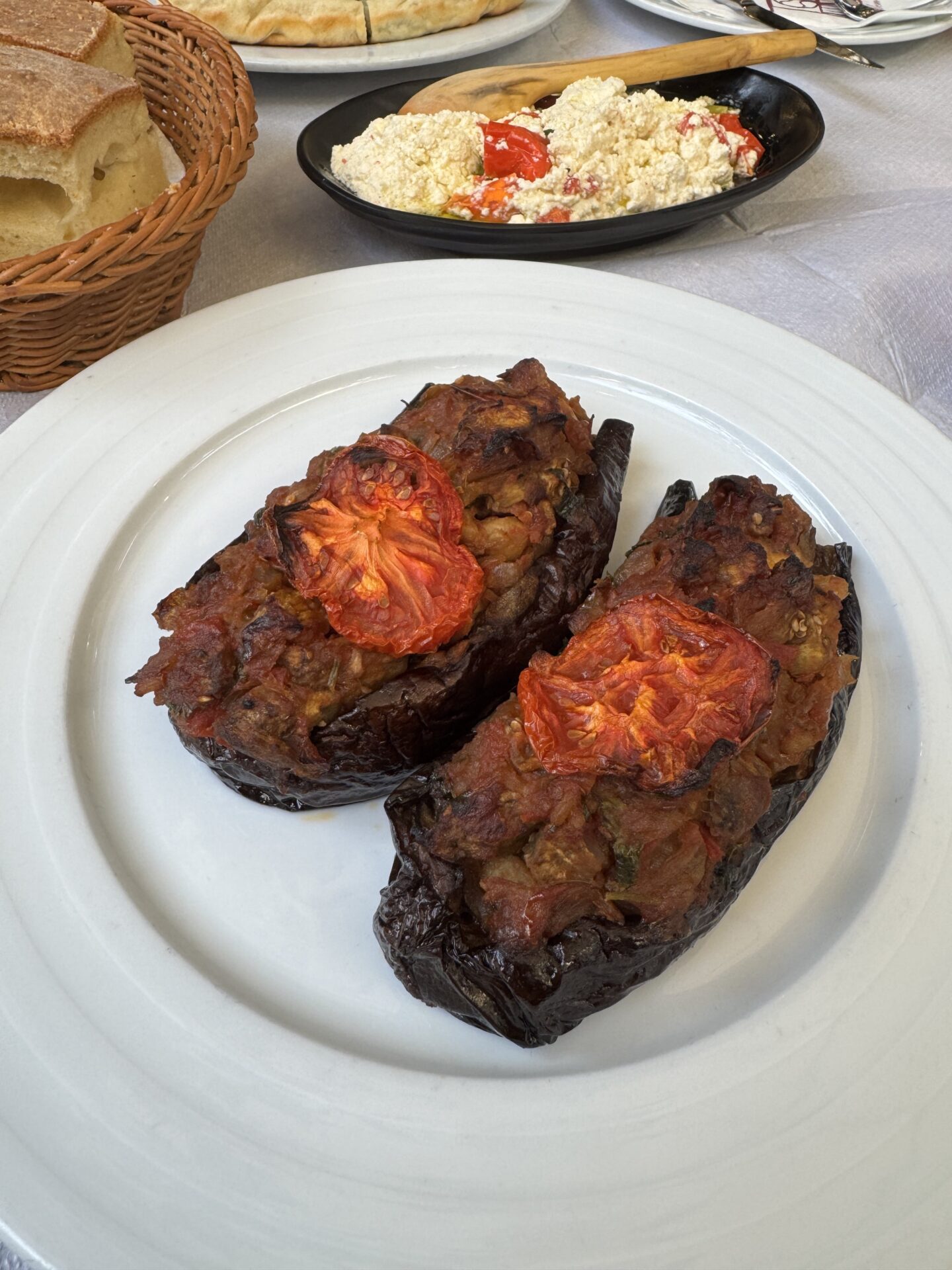 Close-up of Patëllxhanë të mbushur me perime, a traditional Albanian dish of stuffed aubergines. The aubergine is filled with a savoury vegetable mixture and topped with roasted tomato slices, served on a white plate. In the background, there are other dishes, including feta cheese with cherry tomatoes, byrek, and a basket of sliced bread