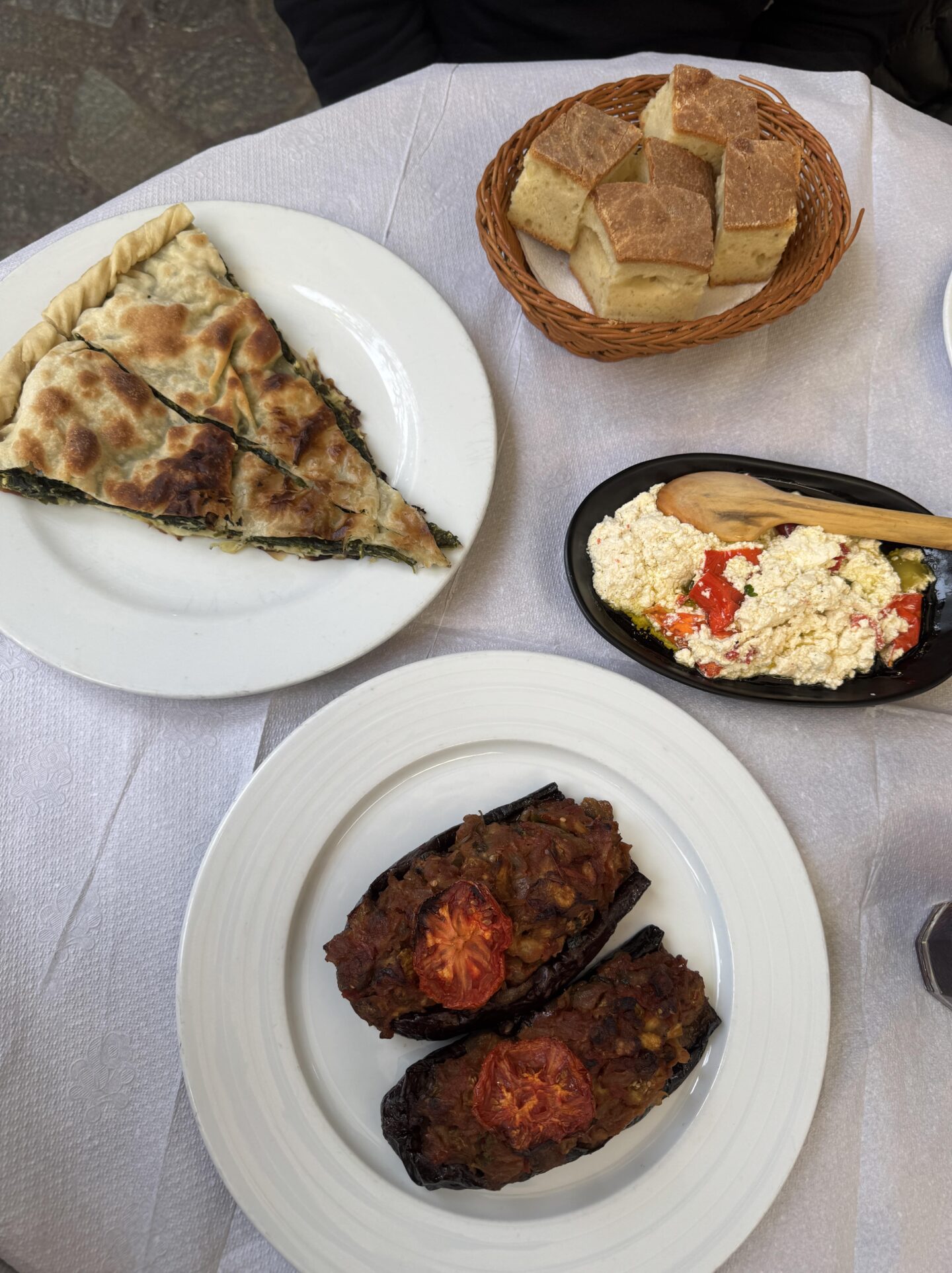 Top-down view of a table with traditional Albanian dishes. The spread includes a slice of lakror (a savoury pastry filled with spinach) on a white plate, Patëllxhanë të mbushur me perime (stuffed aubergines topped with roasted tomatoes), a black dish of feta cheese mixed with peppers and olive oil with a wooden spoon, and a basket of sliced bread. The table is set with white linens and cutlery, creating a rustic and inviting presentation