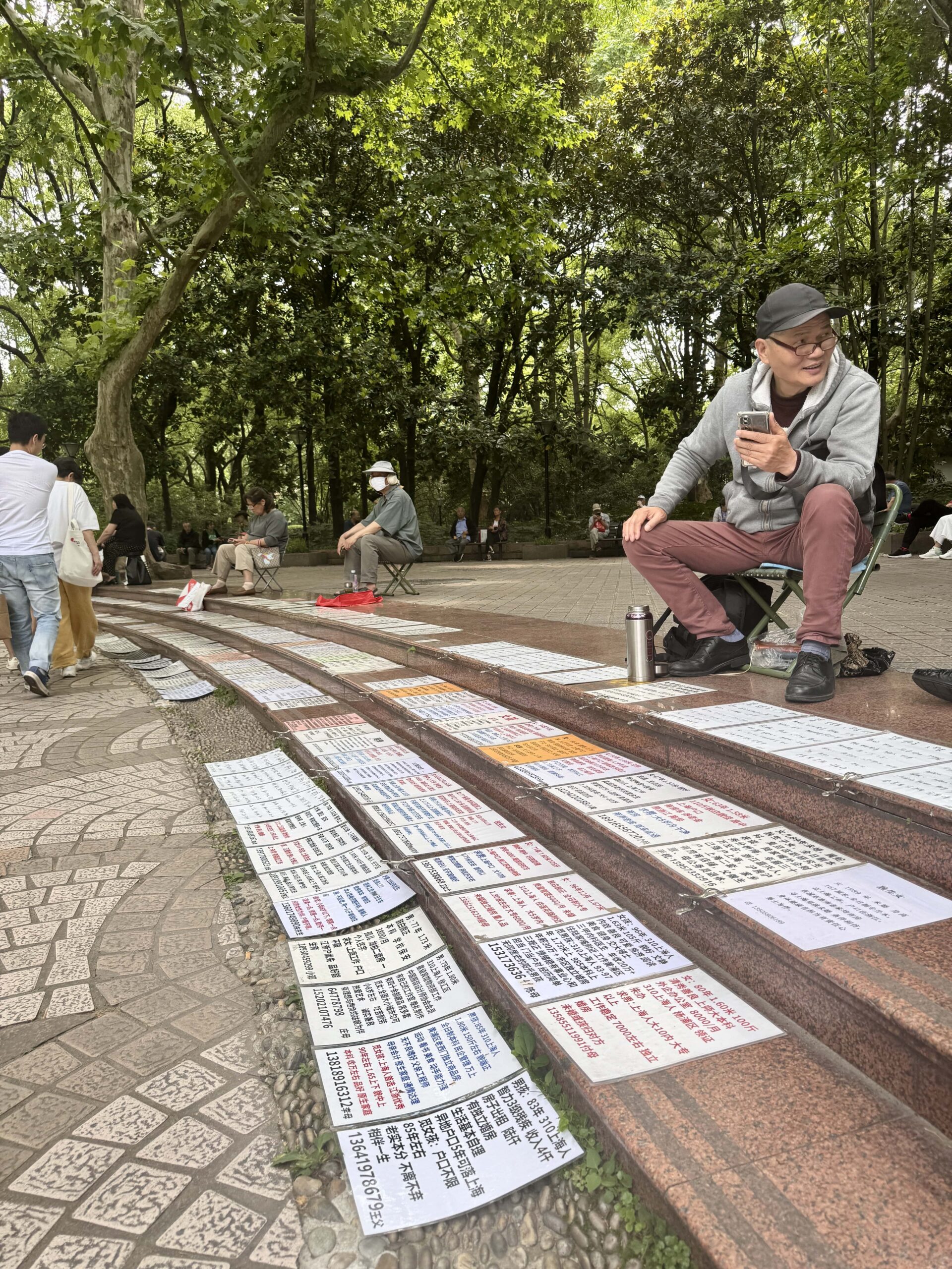 Man sat on a low seat at the top of some steps with rows of resumes in front of him at the Shanghai Marriage Market