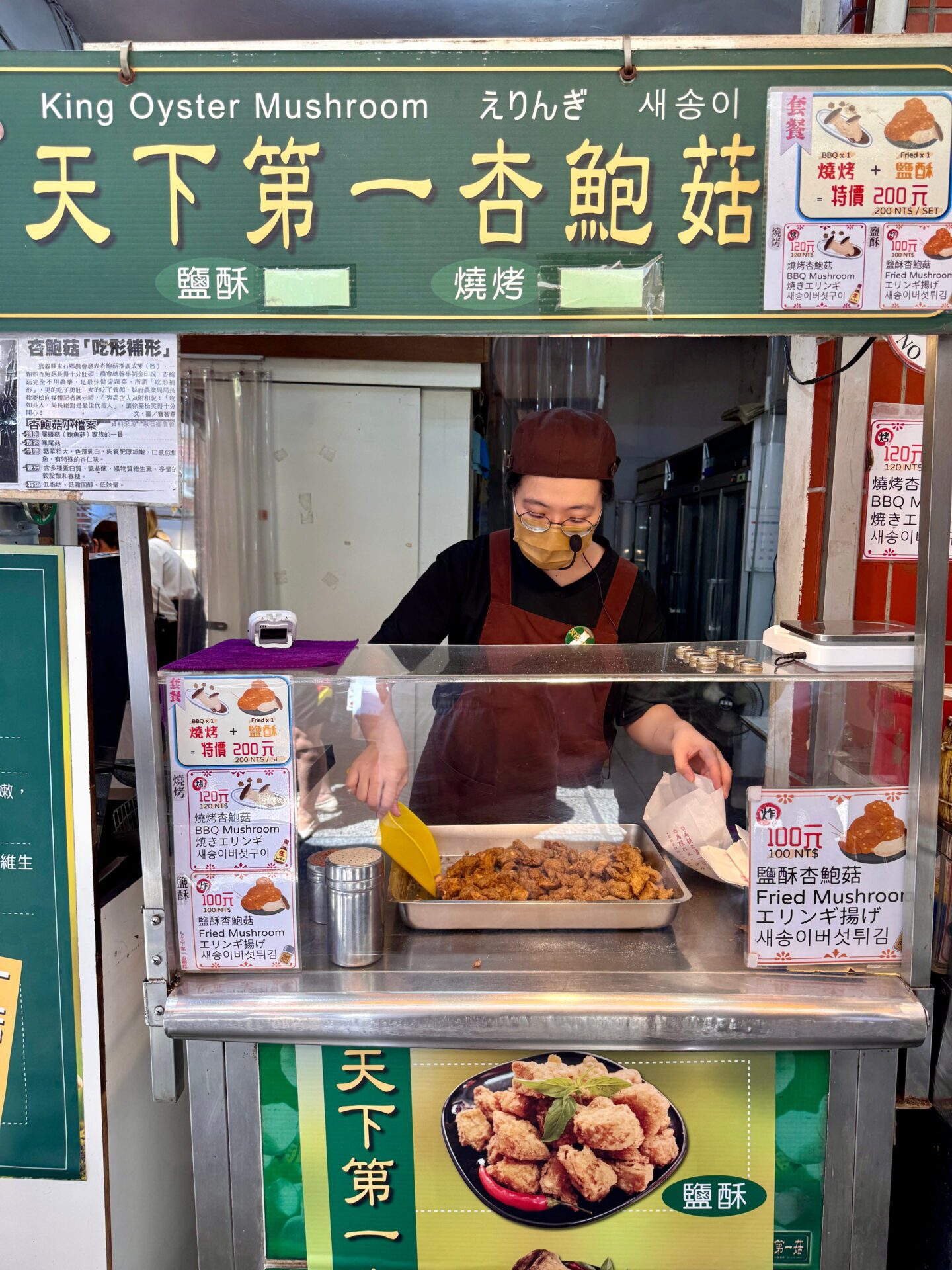 Street food vendor prepares fried king oyster mushrooms behind a glass stall with signs reading King Oyster Mushroom and prices in Chinese and English. A tray of golden brown mushrooms sits in front of the cook, highlighting a popular Taiwanese market snack.