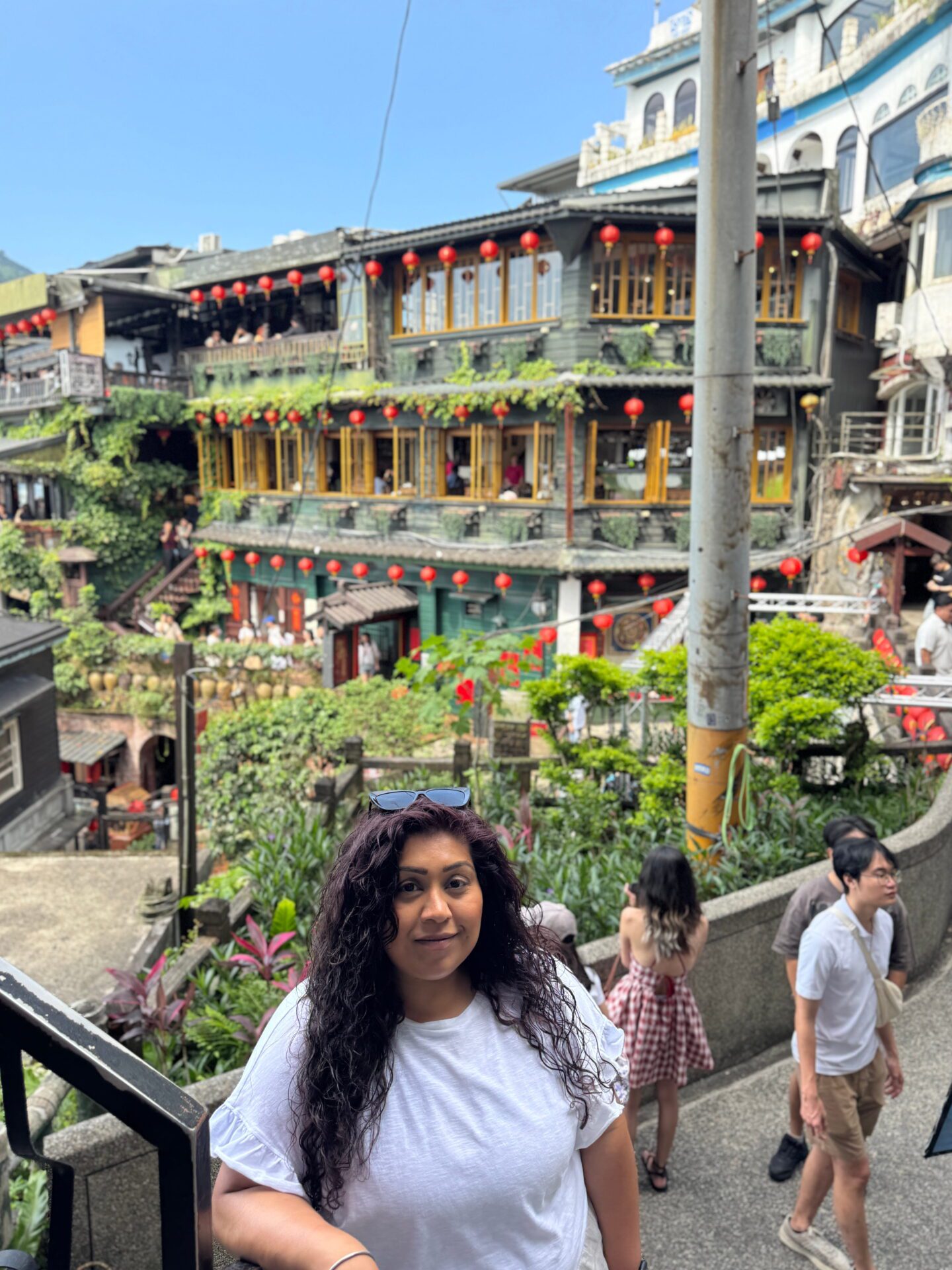 Woman with long curly hair stands on a stone stairway overlooking a hillside of teahouses covered in greenery and lined with red lanterns. The multi level wooden buildings glow with warm yellow windows against a bright blue sky, capturing the lively atmosphere of a mountain village street.