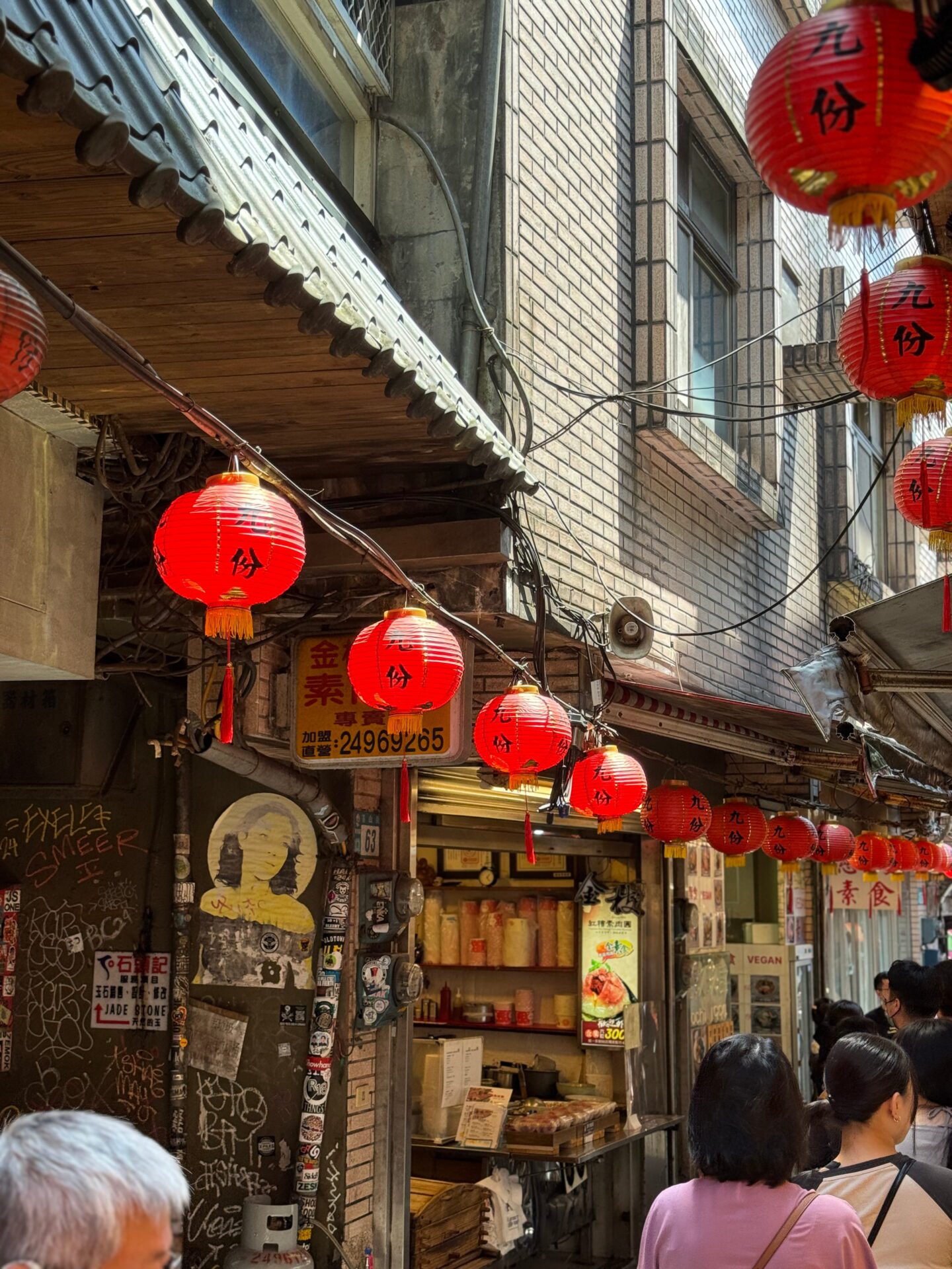 Close view of red lanterns hanging along a brick building above a small street food shop. Signs with Chinese characters and a yellow menu board add to the cozy old town market atmosphere.