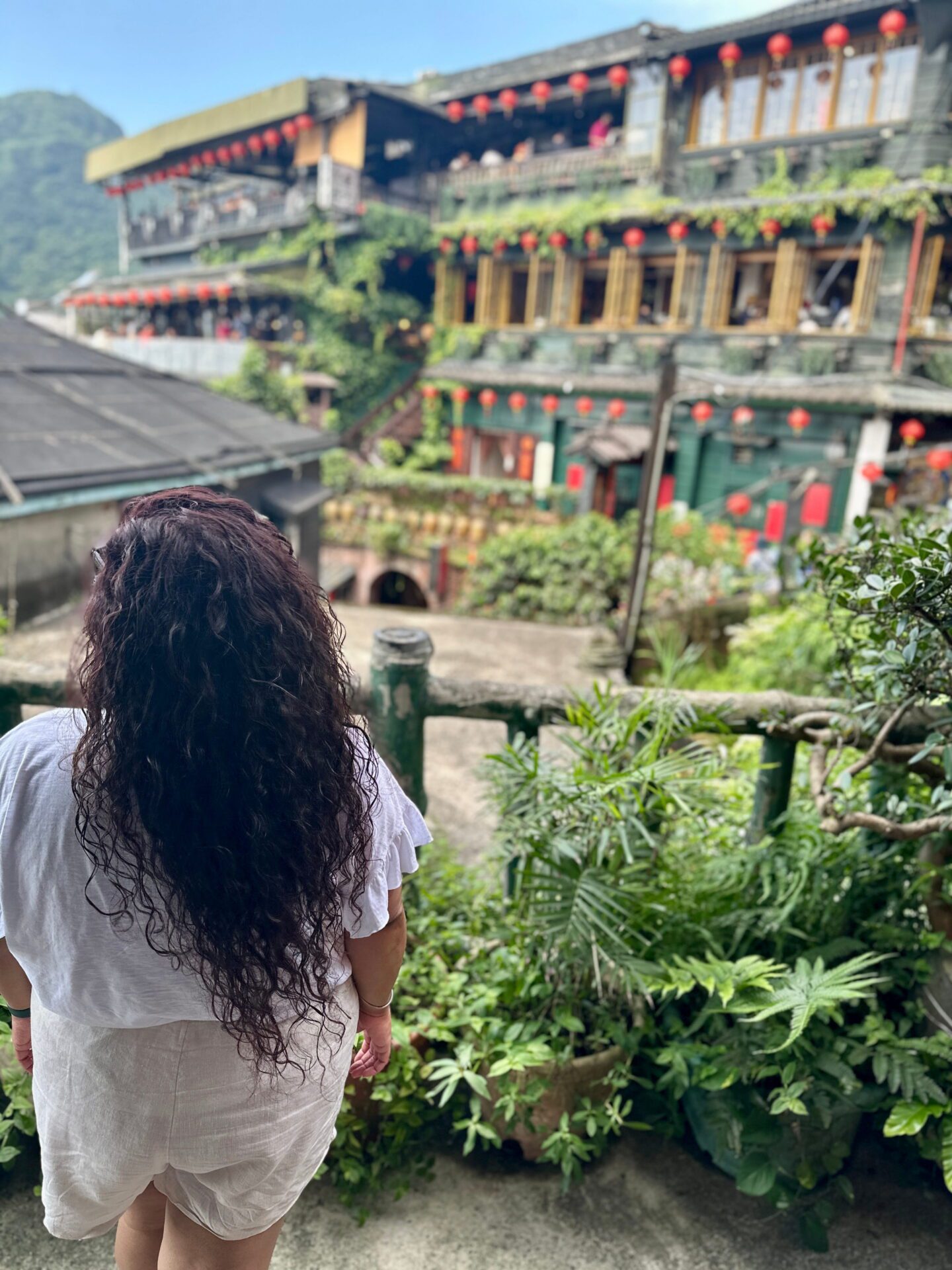 Woman with long curly hair stands facing away from the camera, looking out over lantern covered teahouses surrounded by lush green plants. The layered wooden buildings and mountain backdrop create a scenic hillside village view.