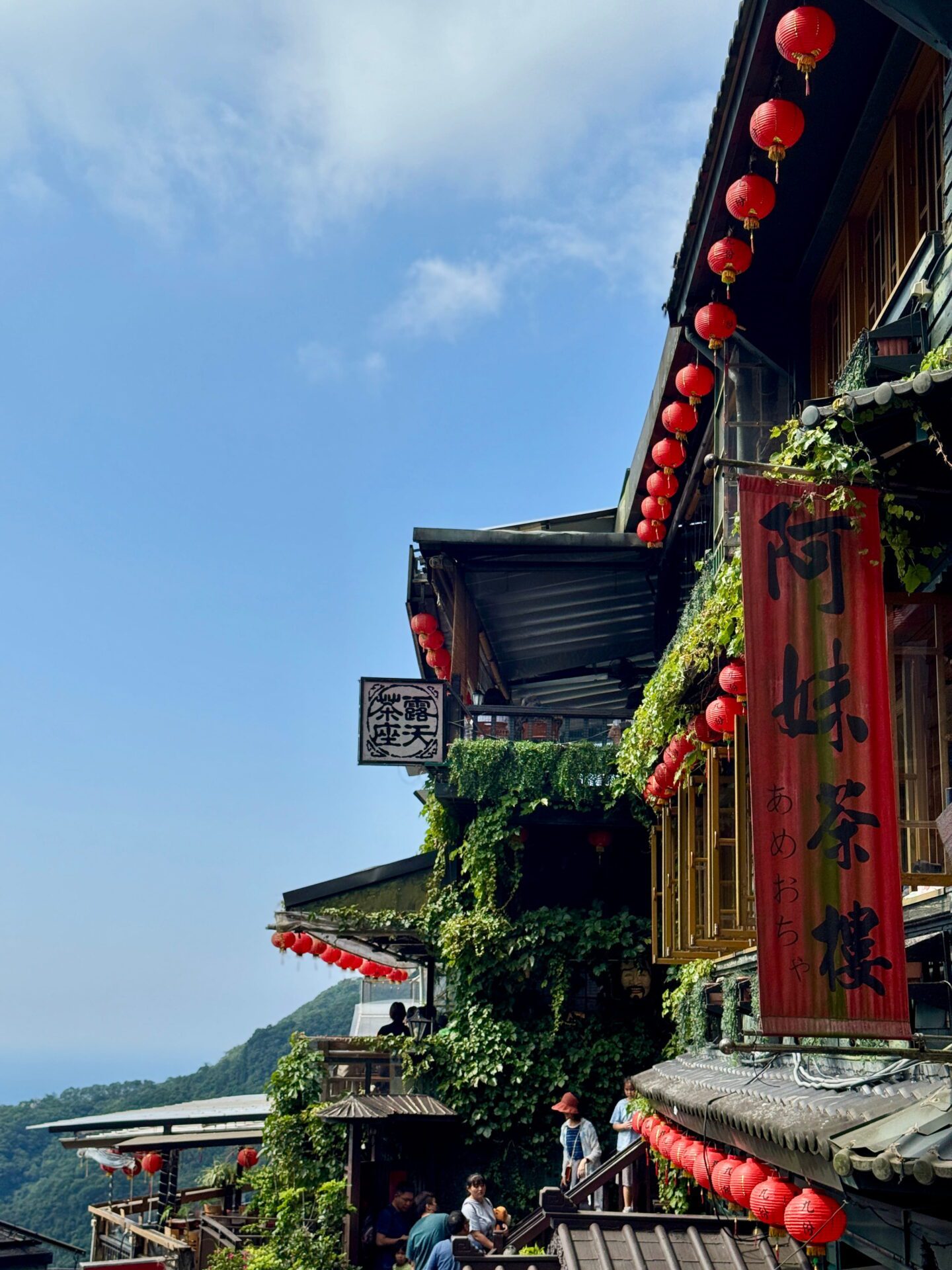 Upward view of a hillside teahouse covered in green vines and lined with rows of red lanterns against a bright blue sky. A vertical red banner with Chinese characters hangs from the building, emphasising the traditional mountain village setting.