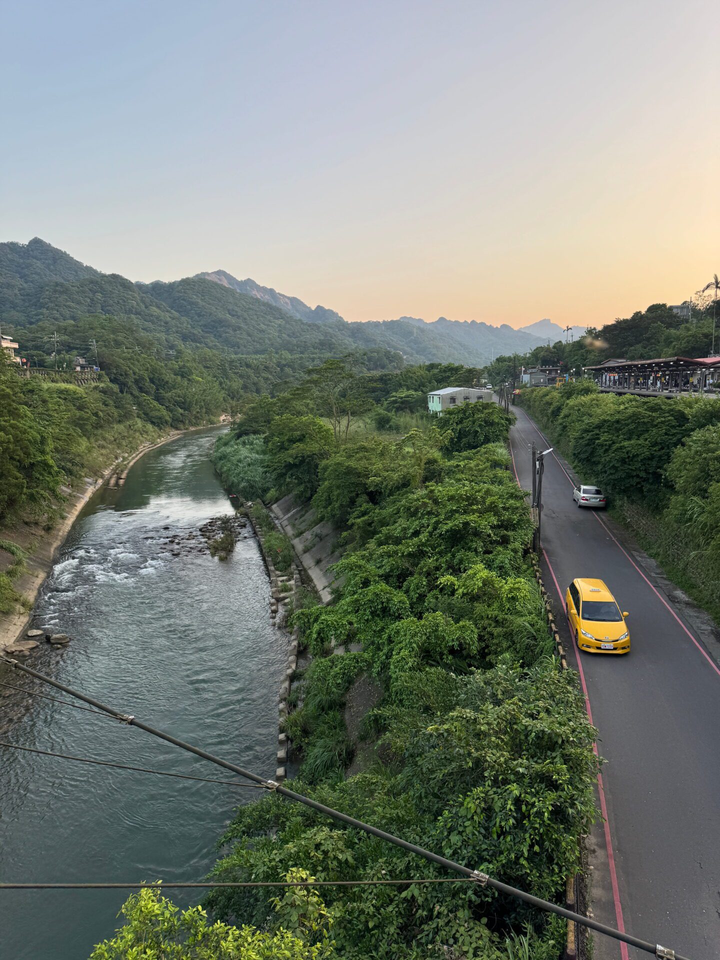 River curves through a lush green valley beside a narrow road with a yellow taxi driving at sunset. Forested hills and a soft orange sky create a peaceful evening landscape.