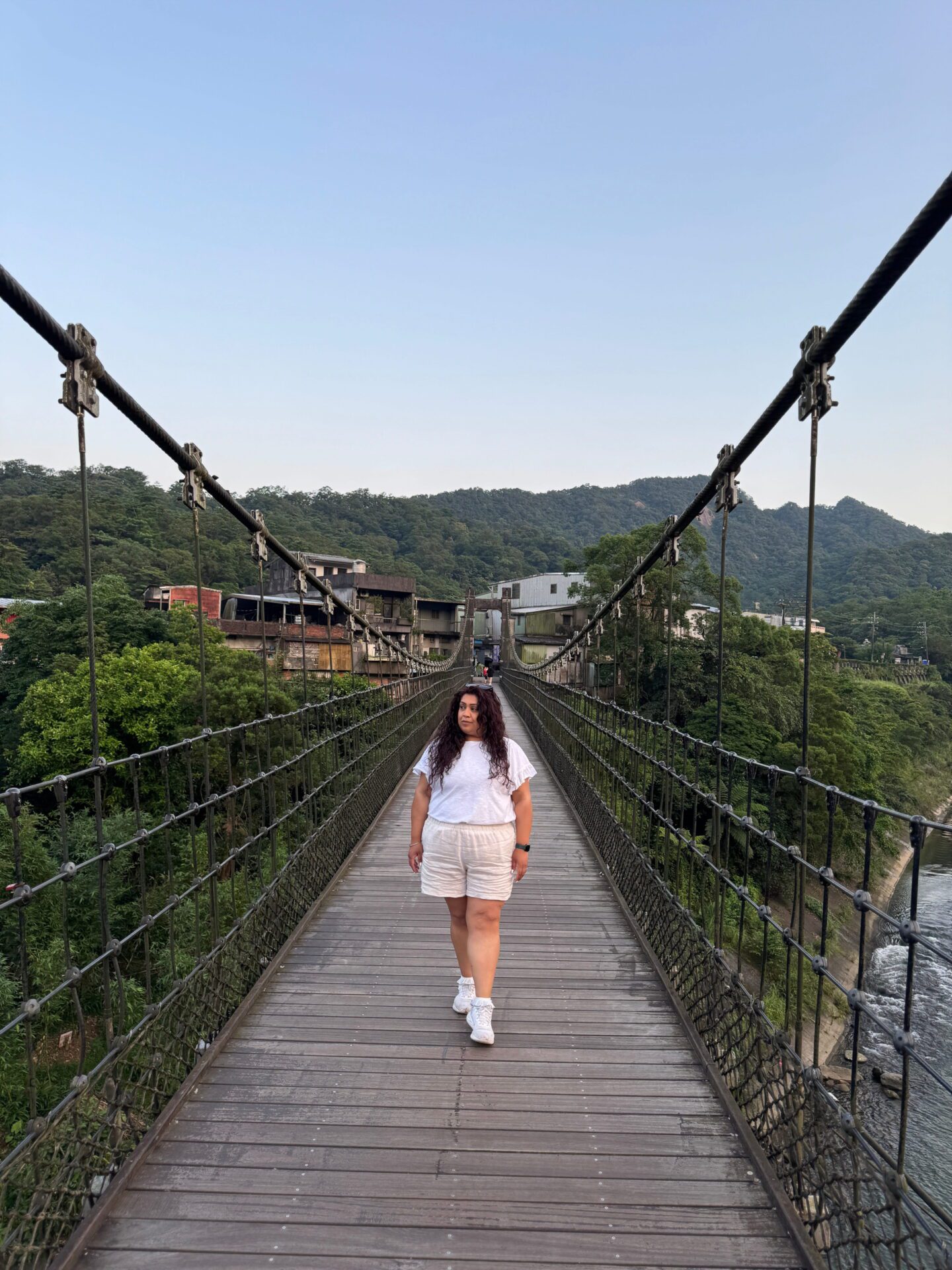 Woman walks along a wooden suspension bridge framed by metal cables and green hills in the distance. The river flows below and low buildings sit at the far end of the bridge.