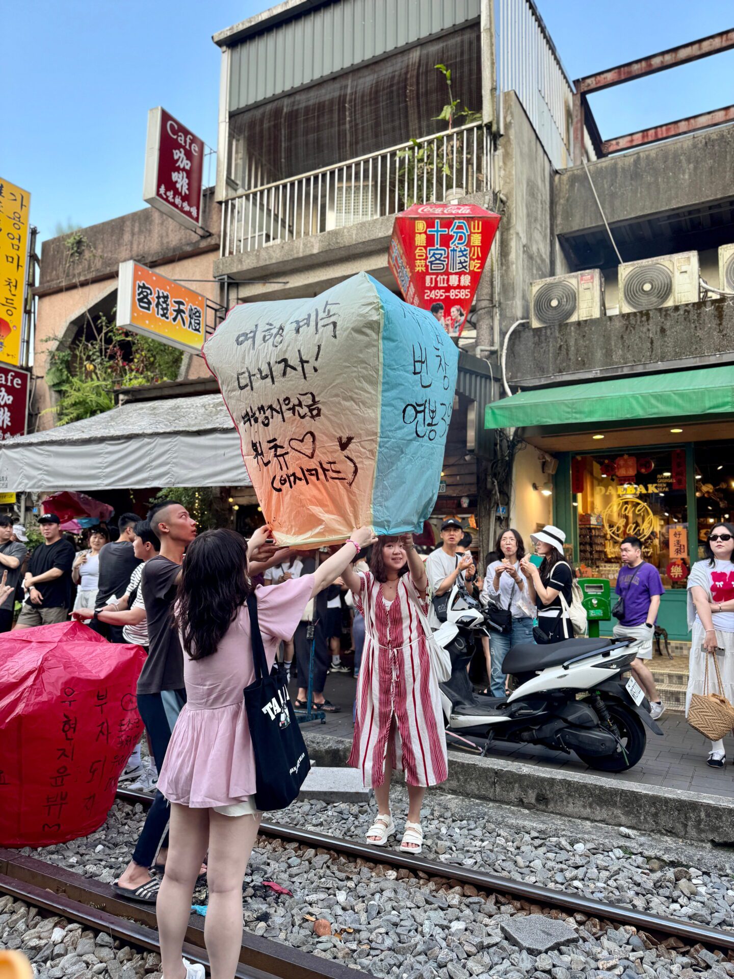 Group of people release colorful sky lanterns over railroad tracks in a busy street scene. The lantern in the center features handwritten Korean messages and small heart drawings, capturing a festive travel moment.