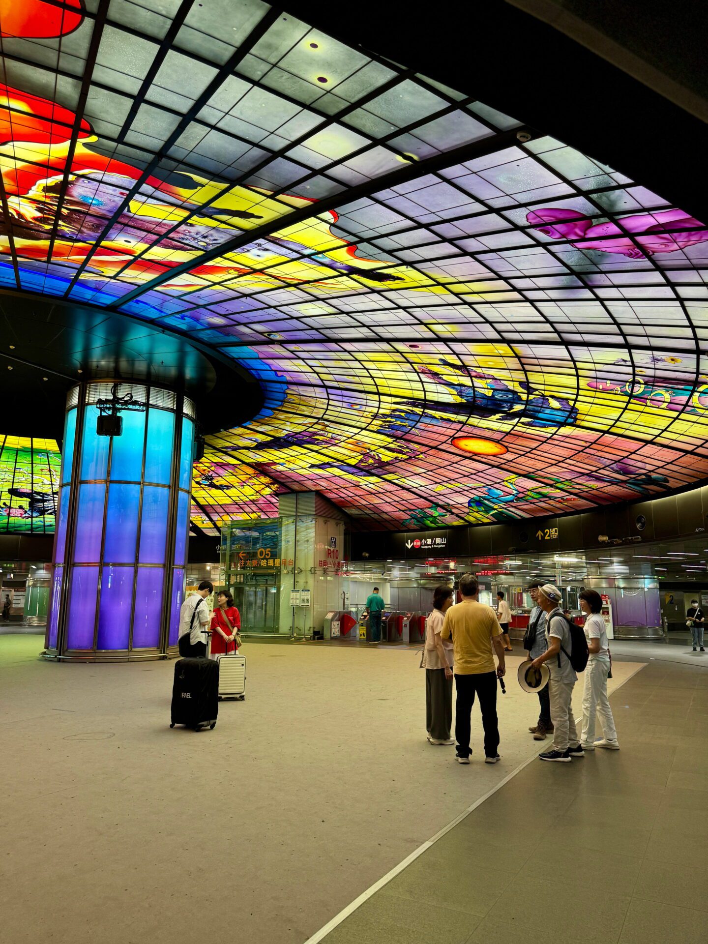 Travellers stand beneath the vivid Dome of Light inside a Kaohsiung metro station surrounded by glowing rainbow columns and intricate glass artwork overhead. This famous installation is a must see when exploring things to do in Kaohsiung.