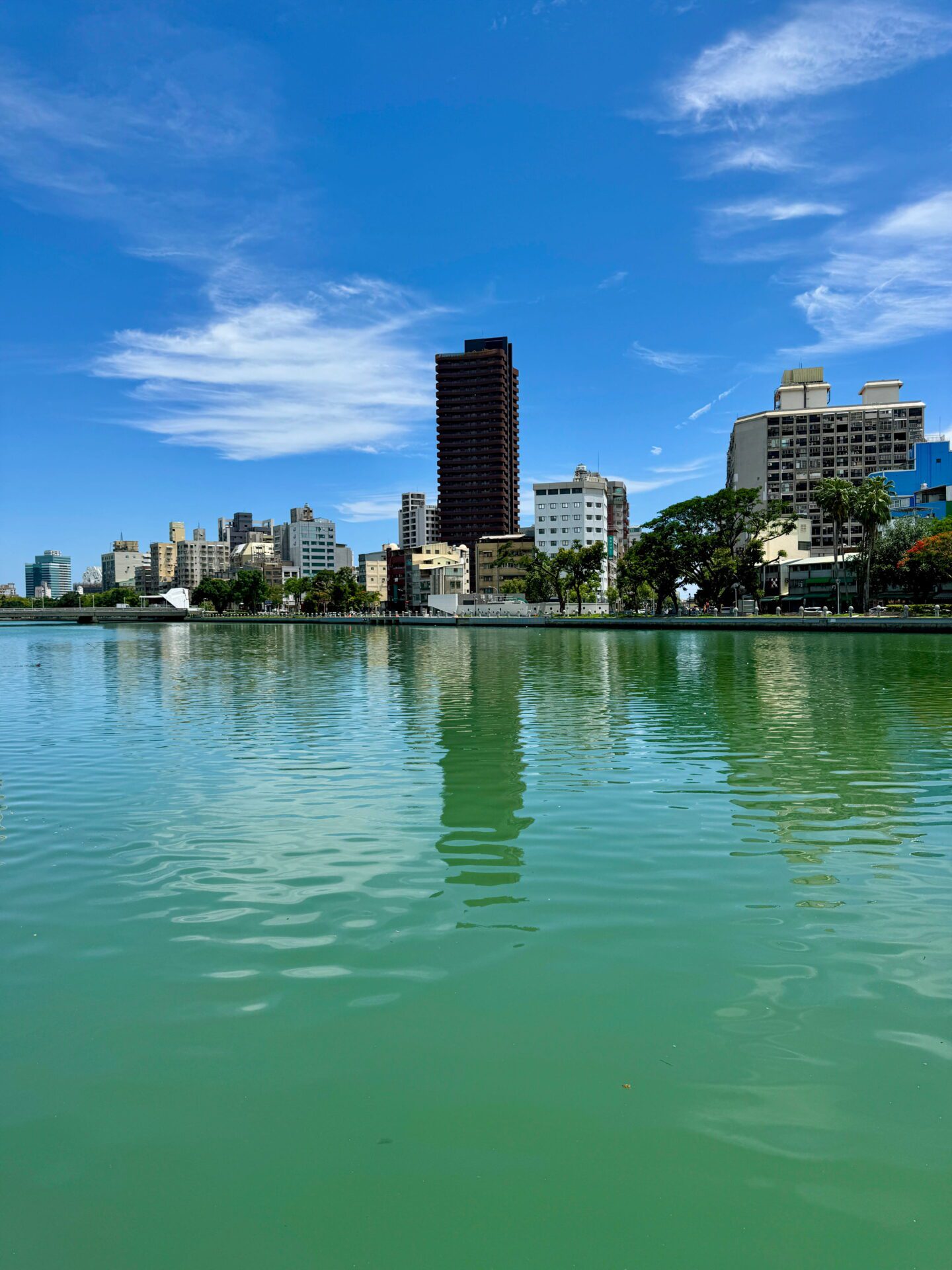 Calm river in Kaohsiung reflecting city buildings under a bright blue sky with scattered clouds and greenery lining the waterfront. A peaceful outdoor scene among relaxing things to do in Kaohsiung.