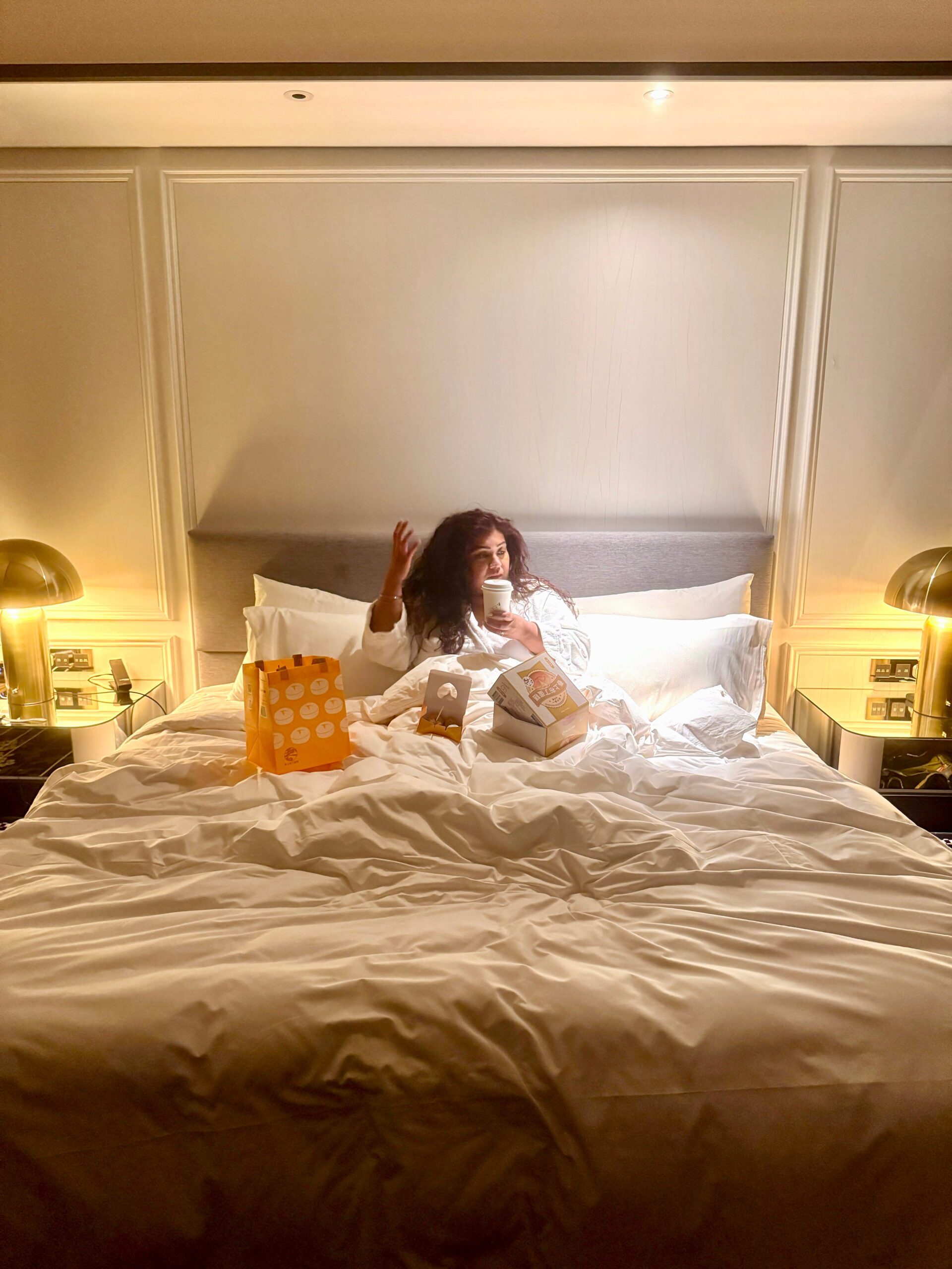 Nishi sitting up in a large hotel bed with white linens and a padded headboard, holding a takeaway coffee cup. On the bed beside her are a bright orange shopping bag and a boxed cake with Chinese text visible on the packaging. Warm bedside lamps on glass nightstands create a cozy evening atmosphere in the elegant room at Le Meridien Taichung