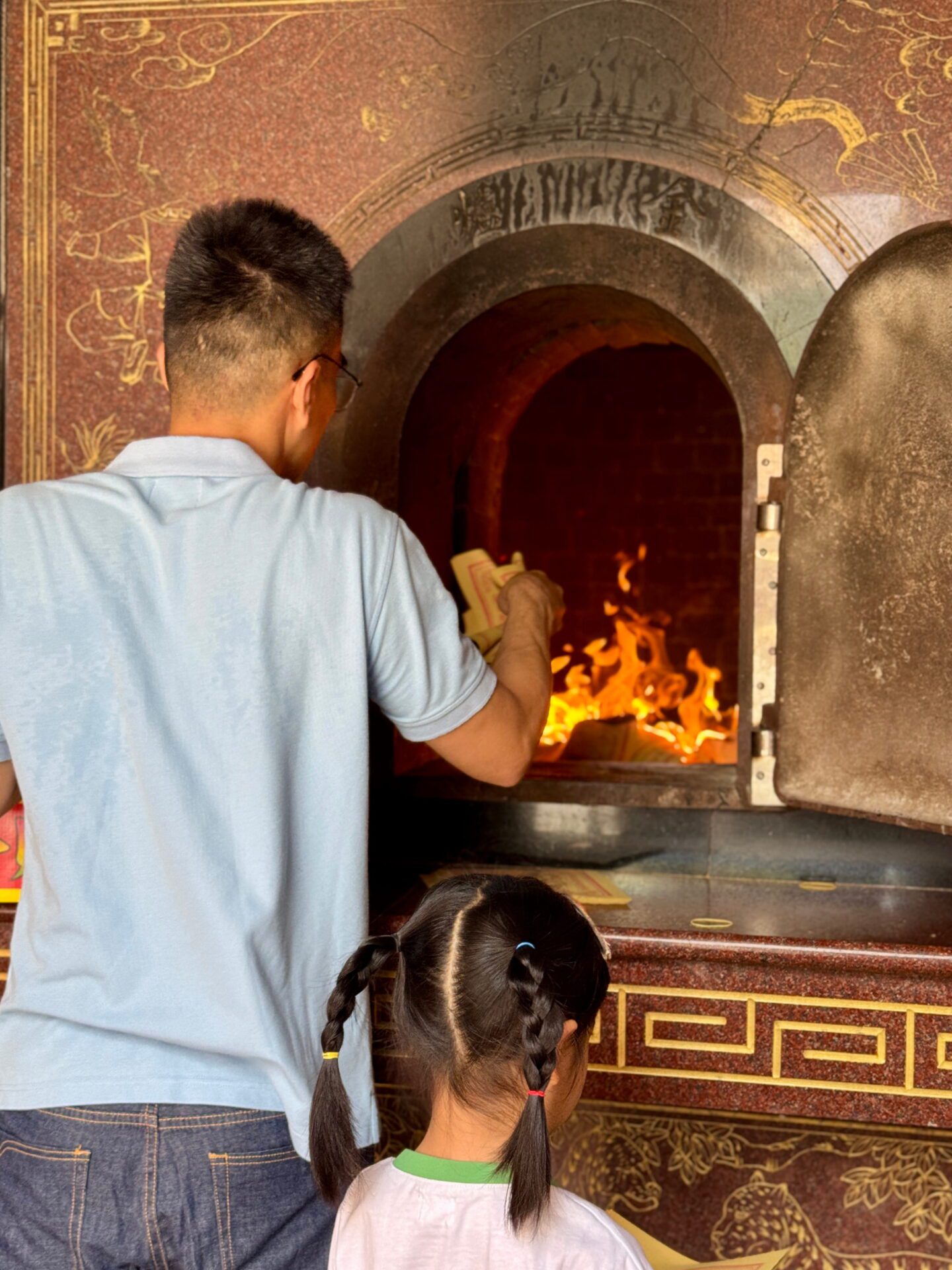 A man and a child place paper offerings into a burning furnace inside Sanfong Temple as flames glow within the arched opening. This ritual is part of local traditions and cultural things to do in Kaohsiung.