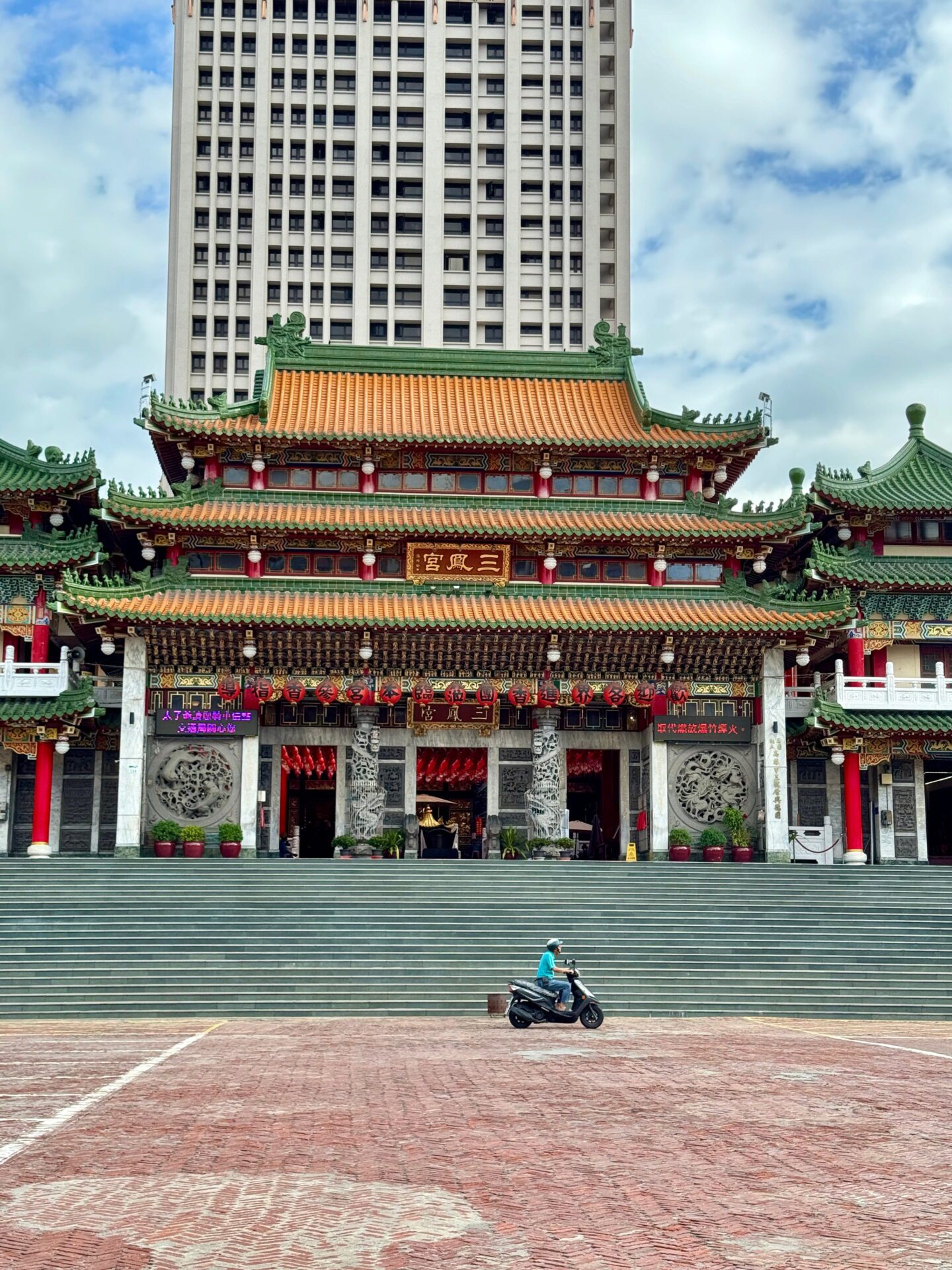 Wide view of Sanfong Temple entrance with grand stairs, detailed carvings, and a scooter passing in front of the building under a cloudy sky. This historic site is a key cultural landmark in Kaohsiung.