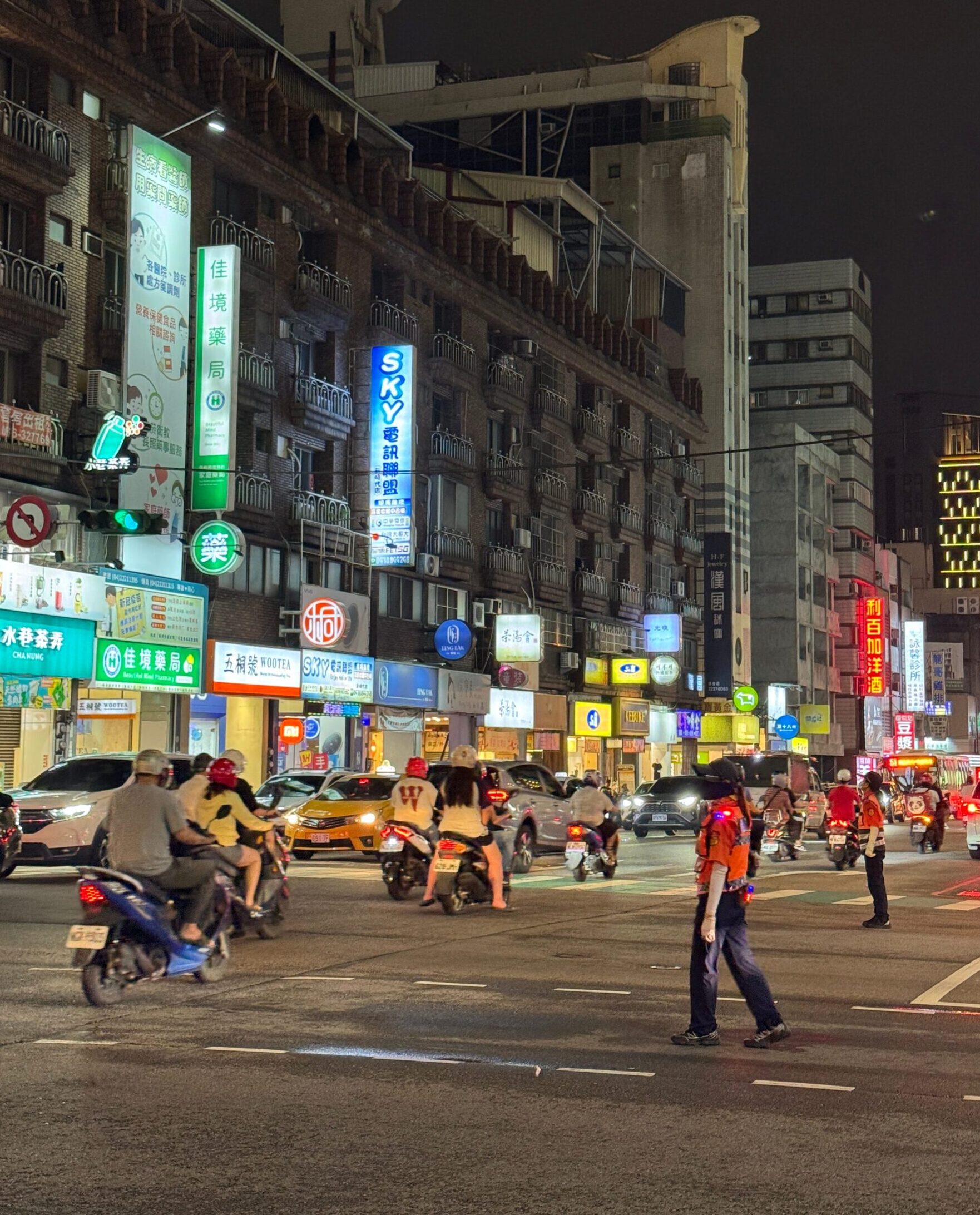 Busy intersection at night filled with scooters and cars beneath bright neon signs and lit shopfronts. A traffic officer in an orange reflective vest stands in the roadway guiding vehicles. The dense signage and glowing lights create a lively urban atmosphere.