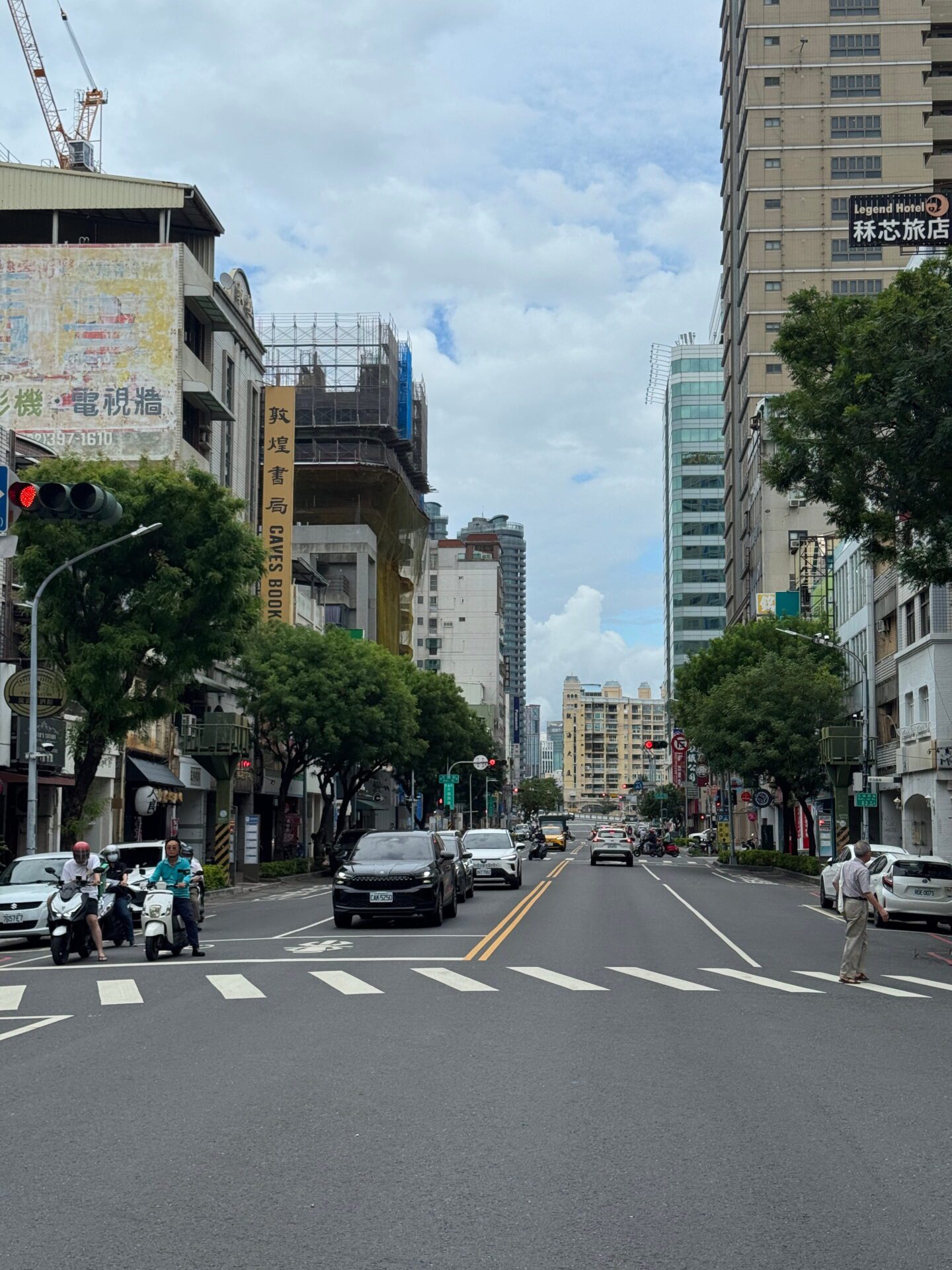 Busy city street in Kaohsiung with cars, scooters, and pedestrians crossing at a junction surrounded by mid rise buildings and shopfront signage.