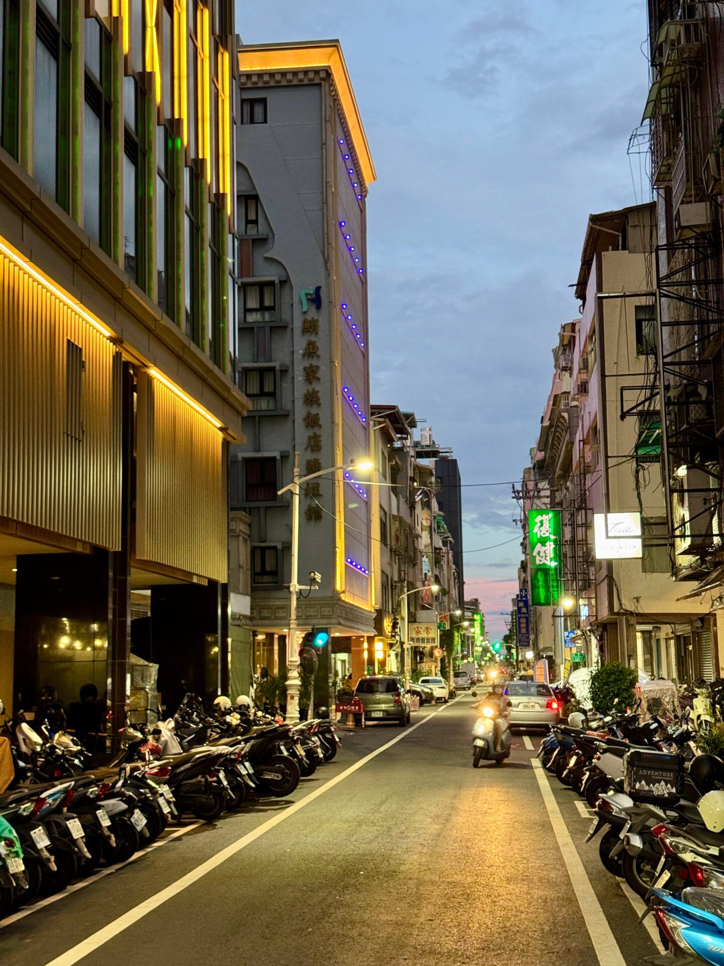 Narrow street in Kaohsiung at night with neon signs, rows of parked scooters, and a motorbike driving through under glowing lights.