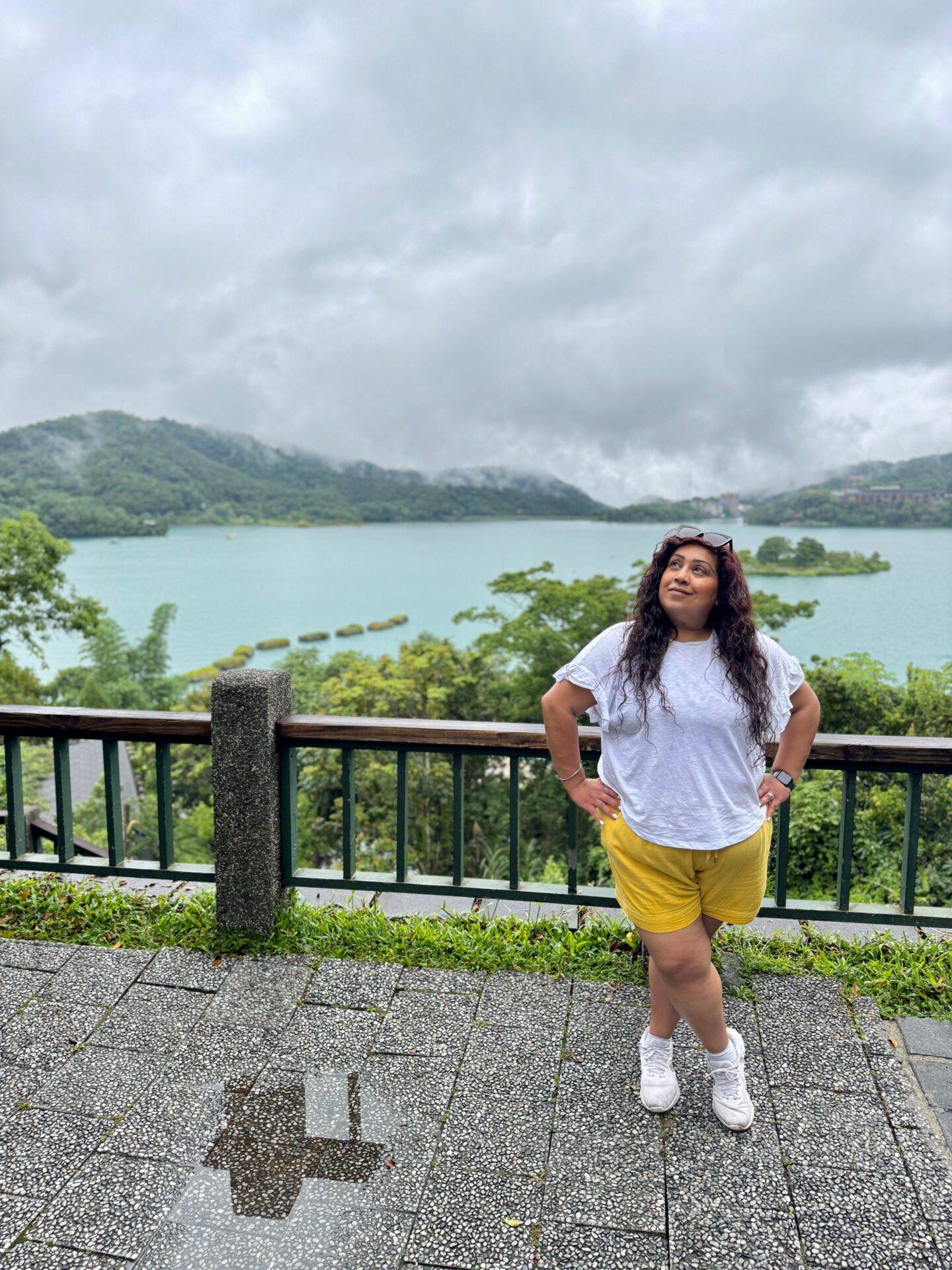 Nishi standing at a scenic viewpoint overlooking Sun Moon Lake with cloudy skies and forested hills surrounding the water. Lalu island and floating barriers appear in the lake behind her.