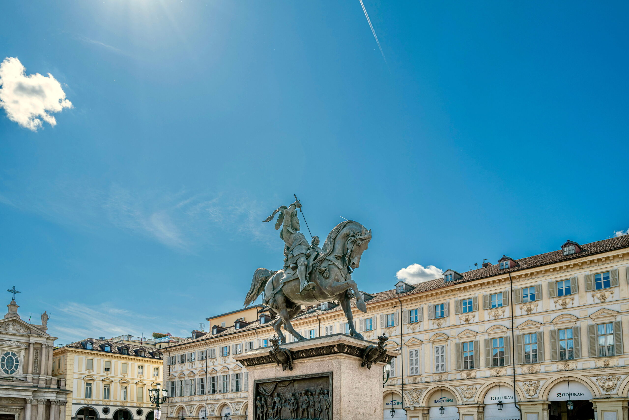 Equestrian statue standing in the centre of the grand Piazza San Carlo Turin square surrounded by ornate cream coloured buildings beneath a vivid blue sky. Bright sunlight highlights the historic architecture and sculpture details.