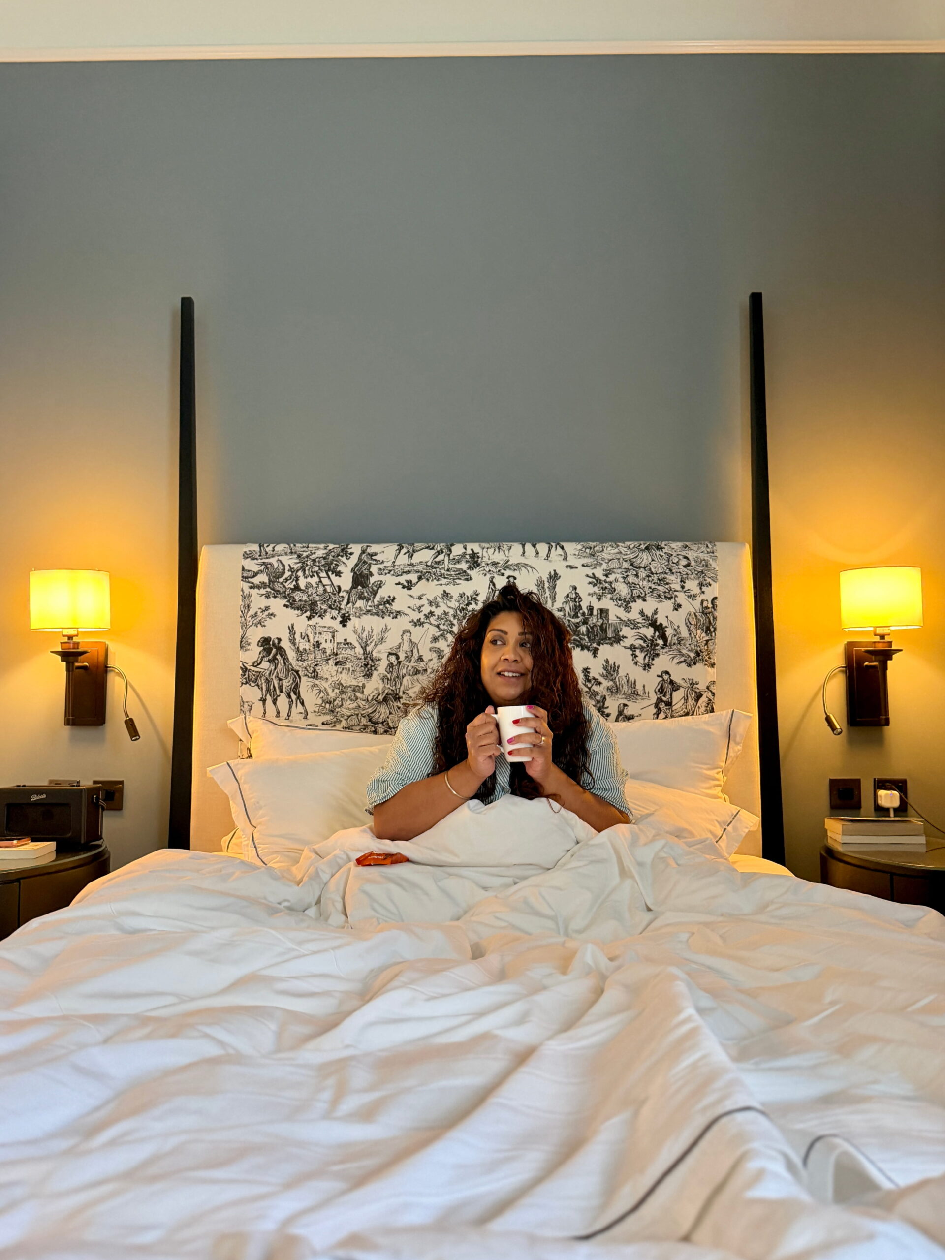 Nishi sitting upright in a large hotel bed holding a hot drink with both hands. Soft bedside lighting and crisp white bedding create a cozy atmosphere for a relaxing morning stay.