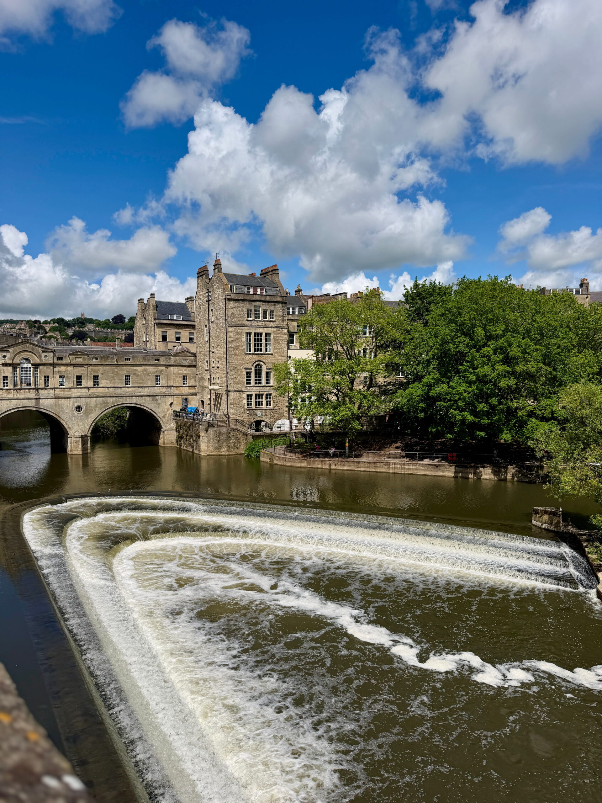 Curved weir on a river flowing beneath a historic stone bridge and buildings under a bright blue sky. White water contrasts with the surrounding greenery and honey coloured architecture.