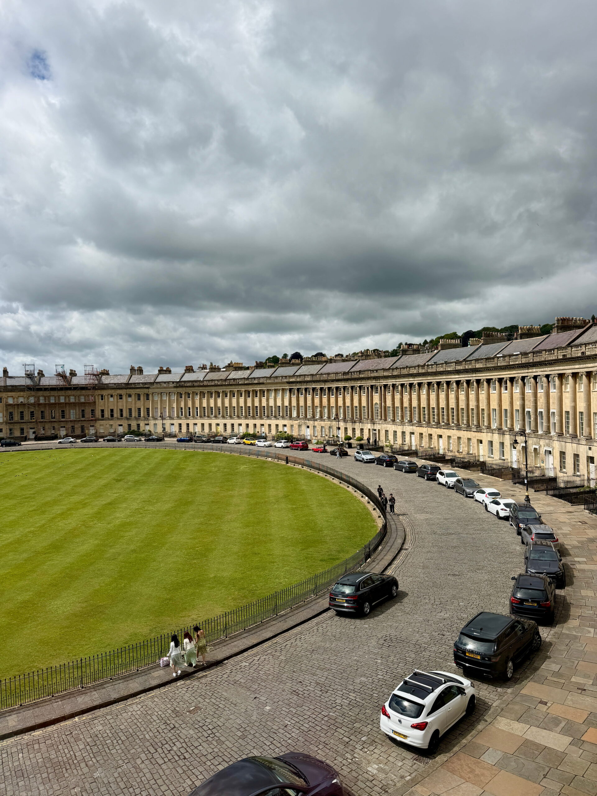 Elevated view of the sweeping crescent of Georgian townhouses surrounding a large green lawn in Bath. Cars line the curved cobblestone road beneath dramatic dark clouds.