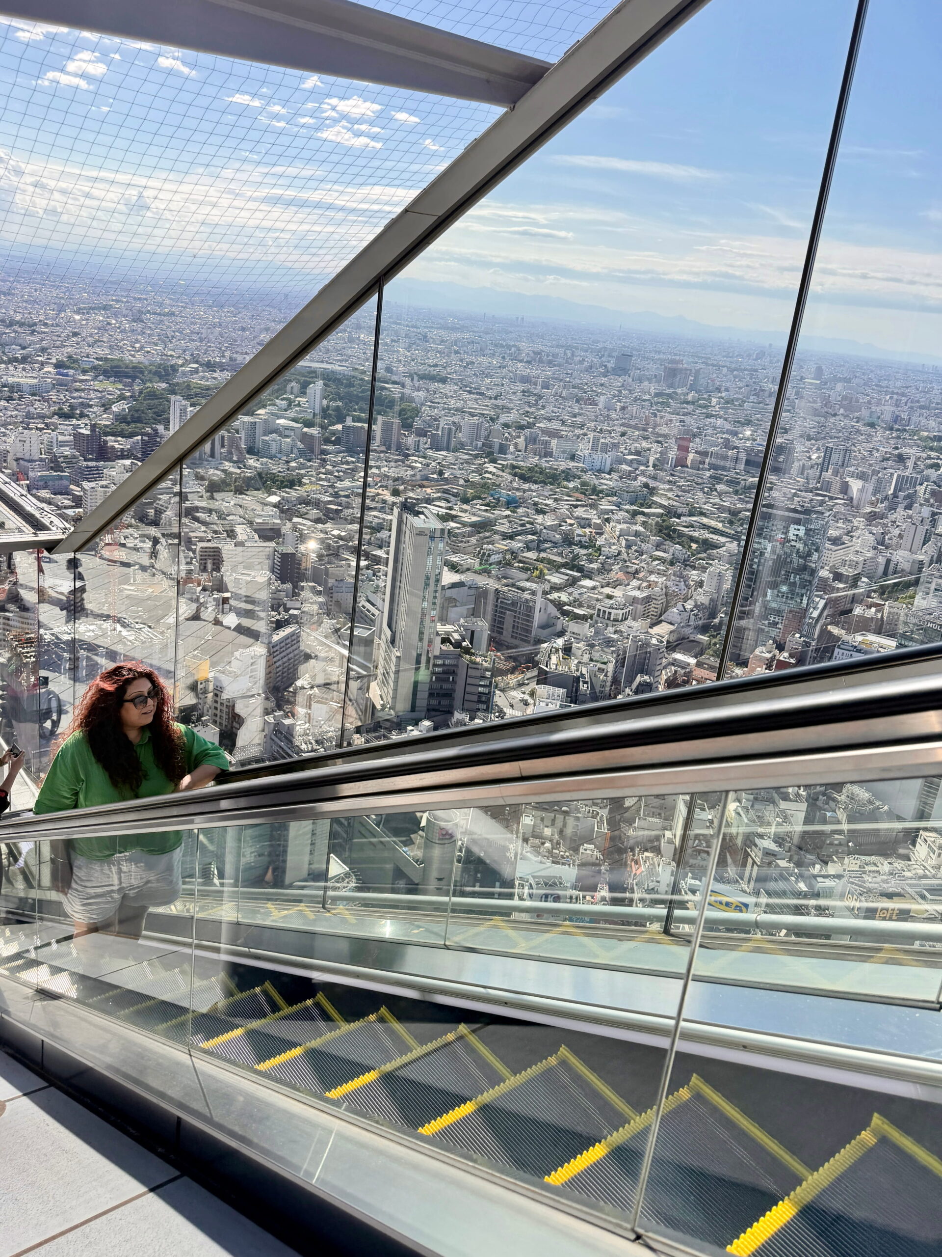 Nishi rides an outdoor escalator high above Tokyo with glass walls revealing the sprawling city far below. The open design at Shibuya Sky creates a dramatic view of the surrounding skyline during the descent.