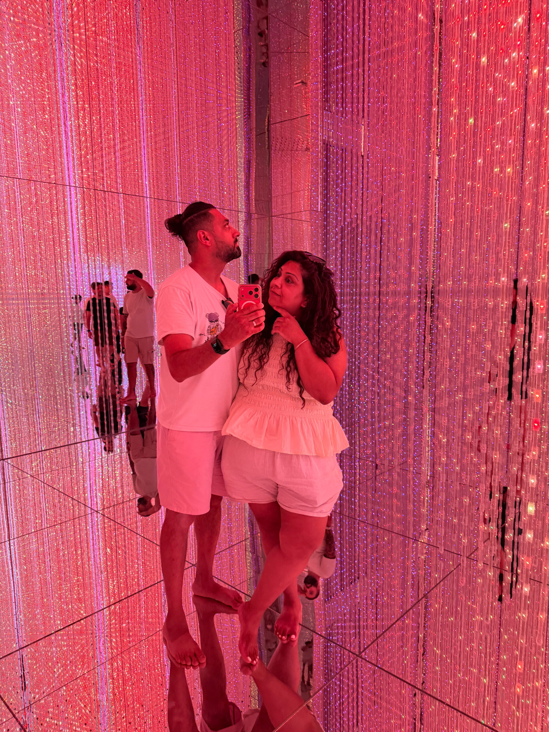 Nishi and Jass taking a mirror selfie surrounded by glowing red and pink strands of light at teamLab Planets Tokyo. The mirrored walls and floor multiply the lights and reflections throughout the immersive room.