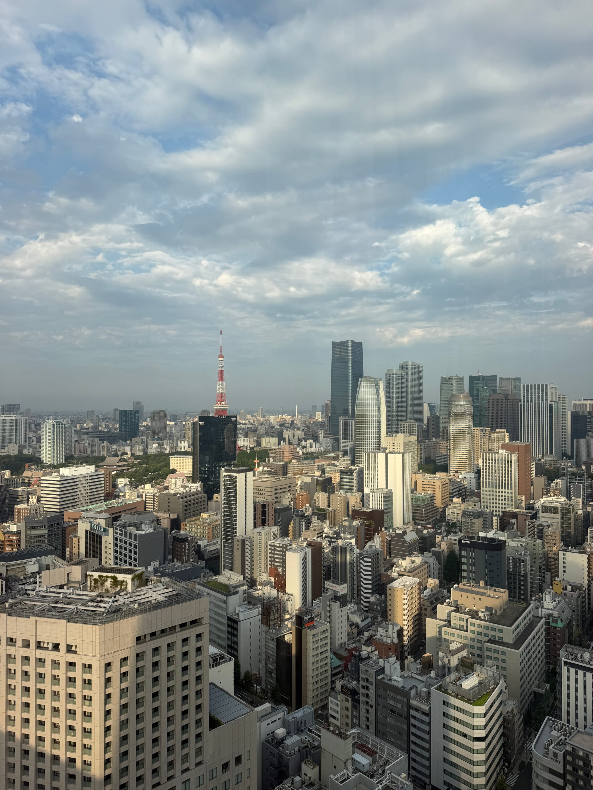 Wide view across the Tokyo skyline from a high floor. Tokyo Tower rises among dense city buildings beneath a partly cloudy blue sky.