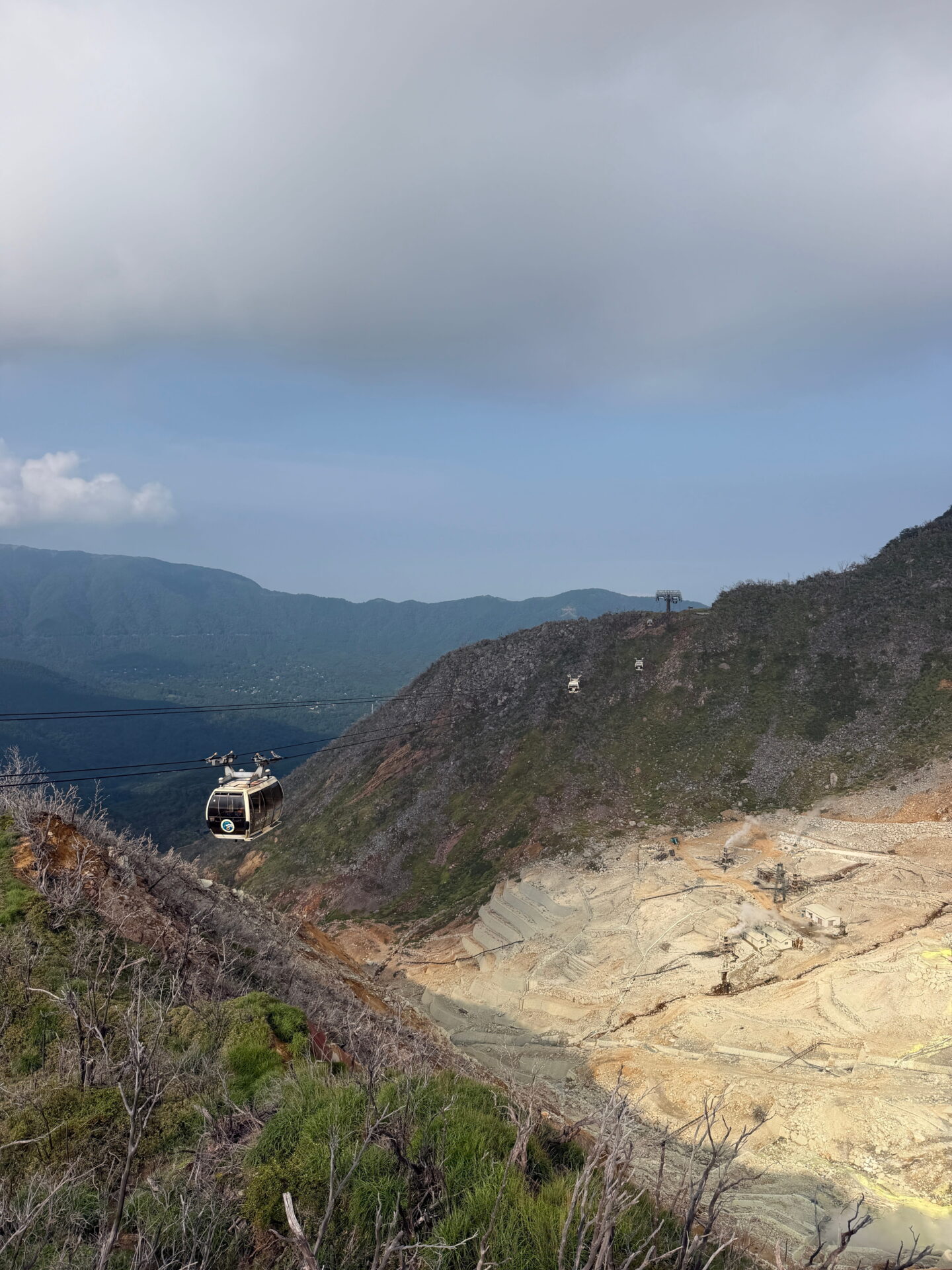 A cable car travels above the steep volcanic landscape towards the station at Owakudani in Hakone. Rocky slopes and rising steam can be seen below beneath a hazy sky.