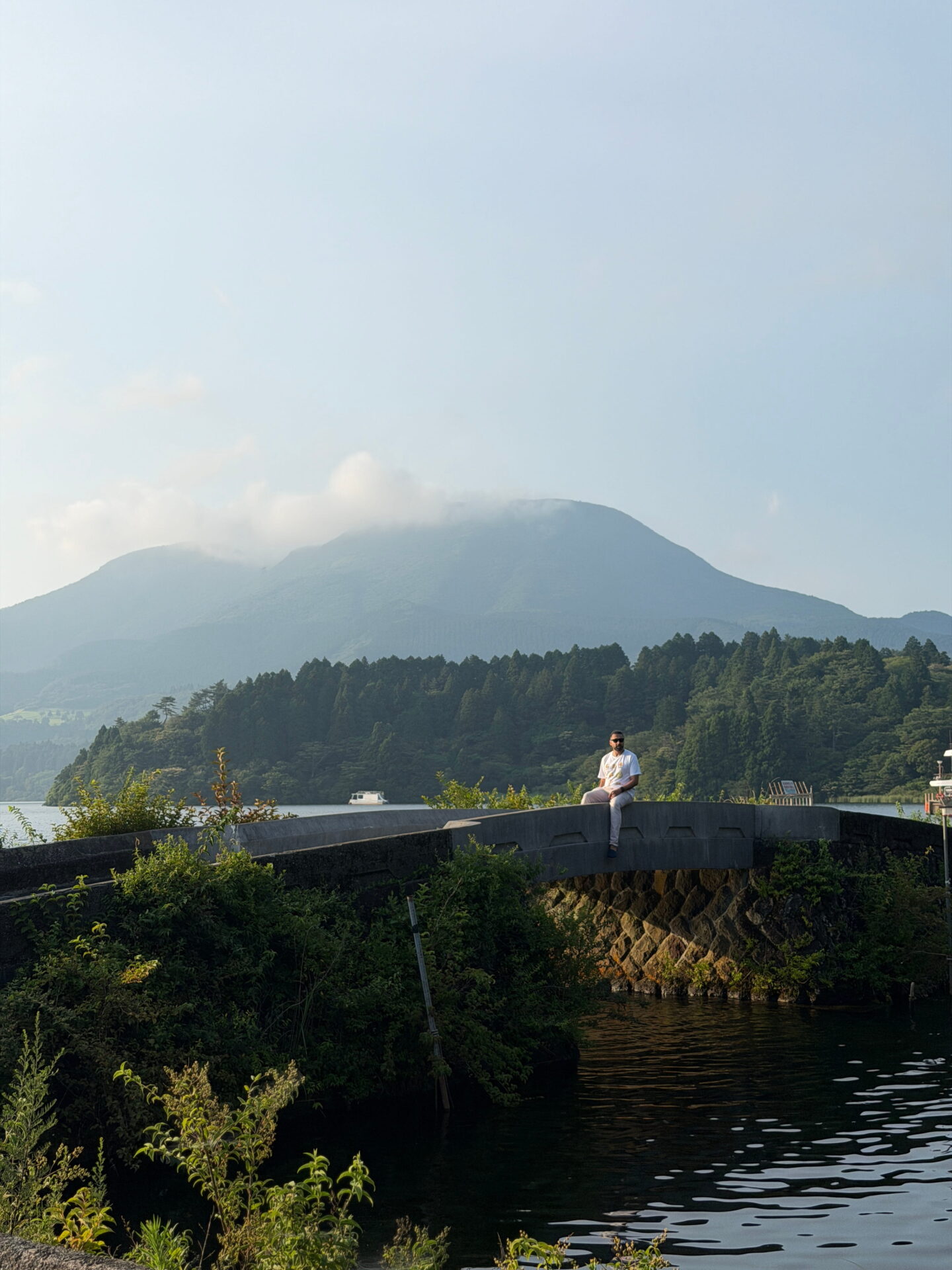 Jass sits on a stone bridge beside Lake Ashi with forested mountains rising behind him in Hakone. Low clouds drift around the mountain peaks as warm evening sunlight falls across the lakeside.