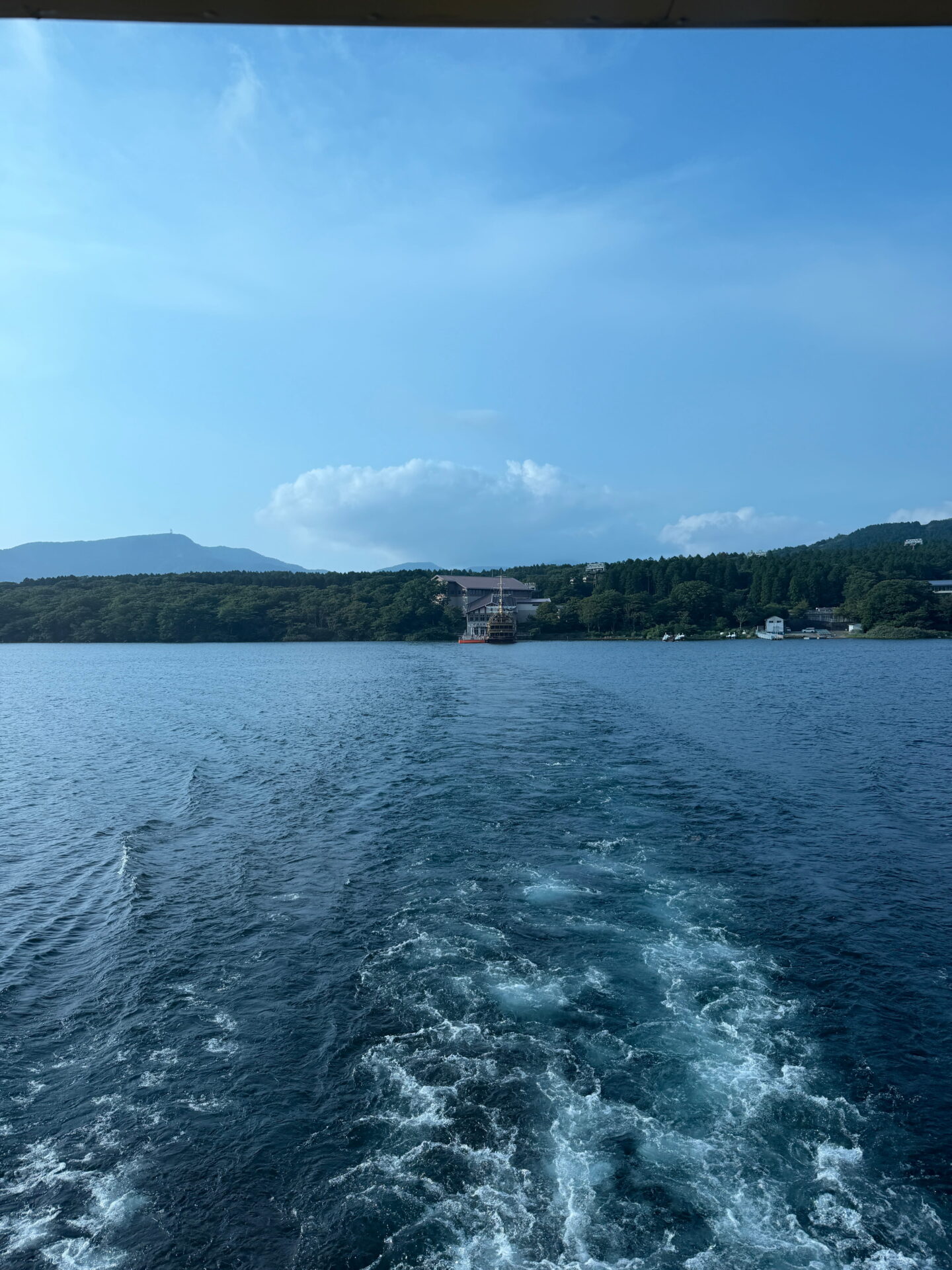 Looking across Lake Ashi from a sightseeing boat in Hakone with its foamy wake trailing through the deep blue water. Forested hills and buildings line the distant shore beneath a bright blue sky.