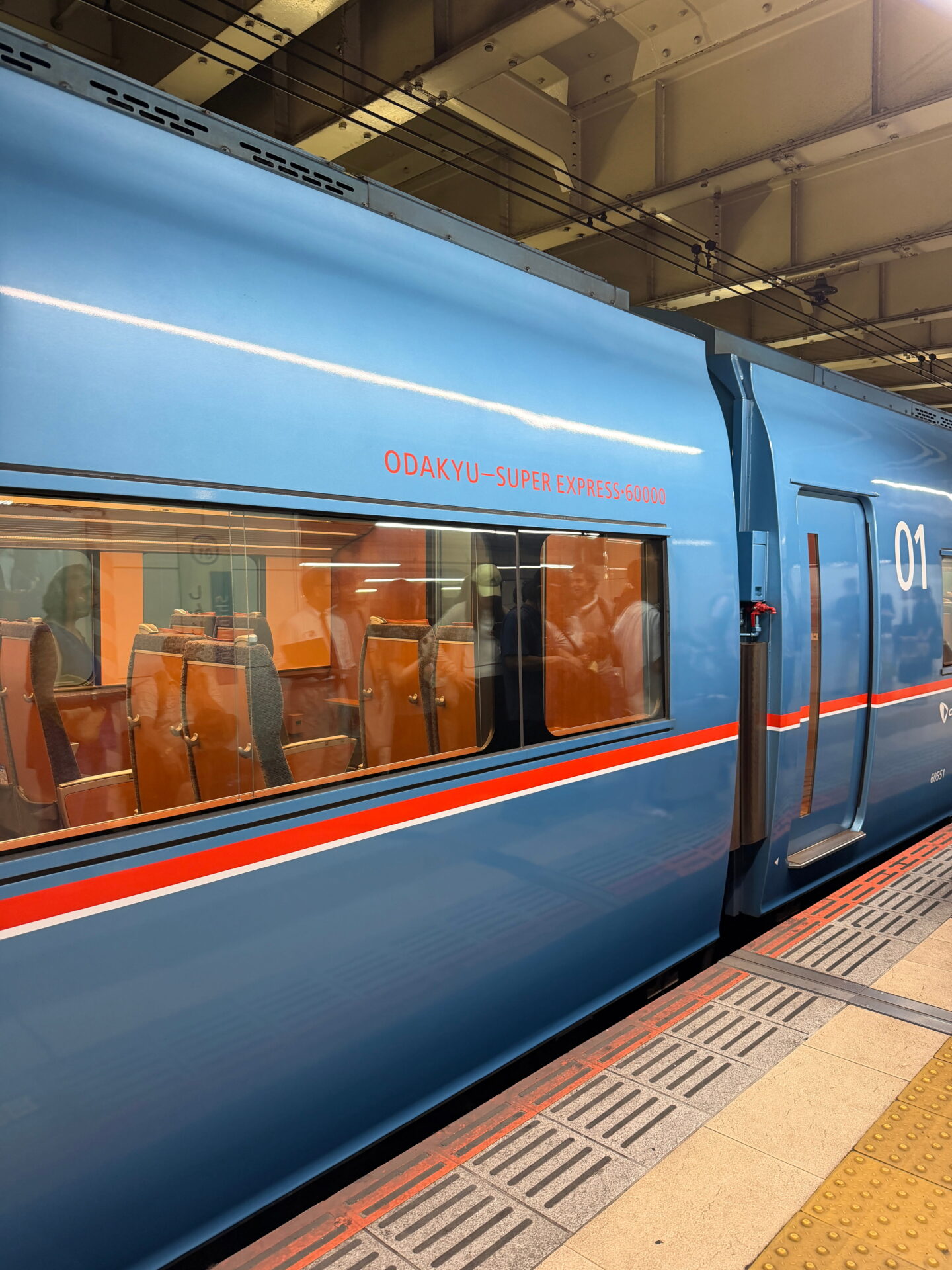Blue Odakyu Romancecar train waiting at a station with large windows showing the passenger seats inside. Red lettering on the side reads "ODAKYU SUPER EXPRESS 60000"