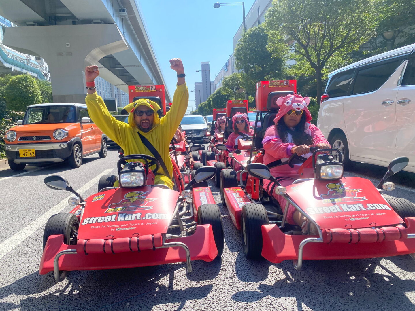 Jass throws both arms into the air while seated in his red Street Kart beside Nishi on a sunny Tokyo road. Jass wears a yellow Pikachu costume while Nishi wears a bright pink character costume and yellow heart shaped sunglasses.