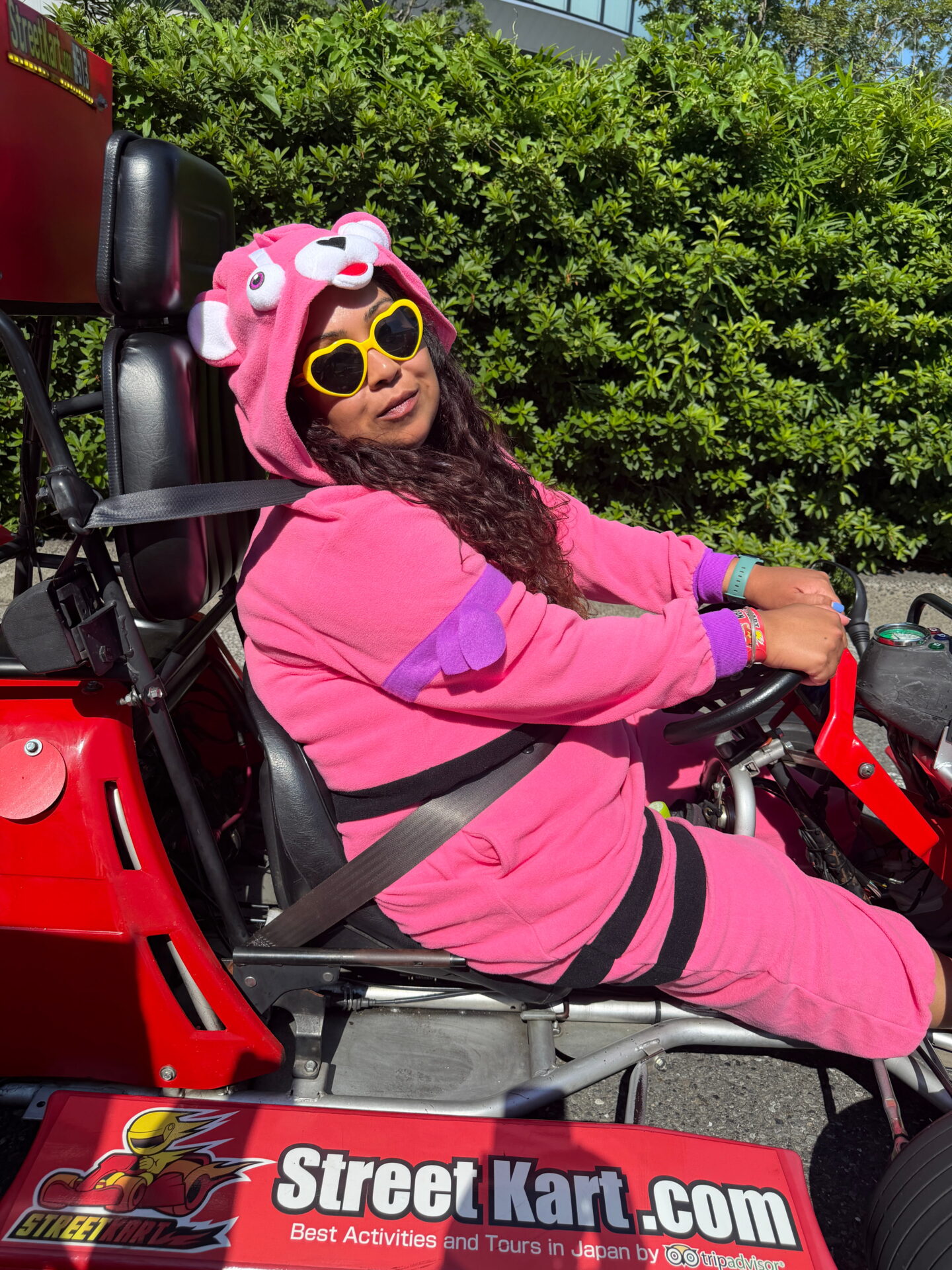 Nishi sits behind the wheel of a red Street Kart wearing a bright pink character costume and yellow heart shaped sunglasses. She is buckled into the kart in the sunshine with green bushes behind her and the side panel reading "StreetKart.com" and "Best Activities and Tours in Japan by Tripadvisor."