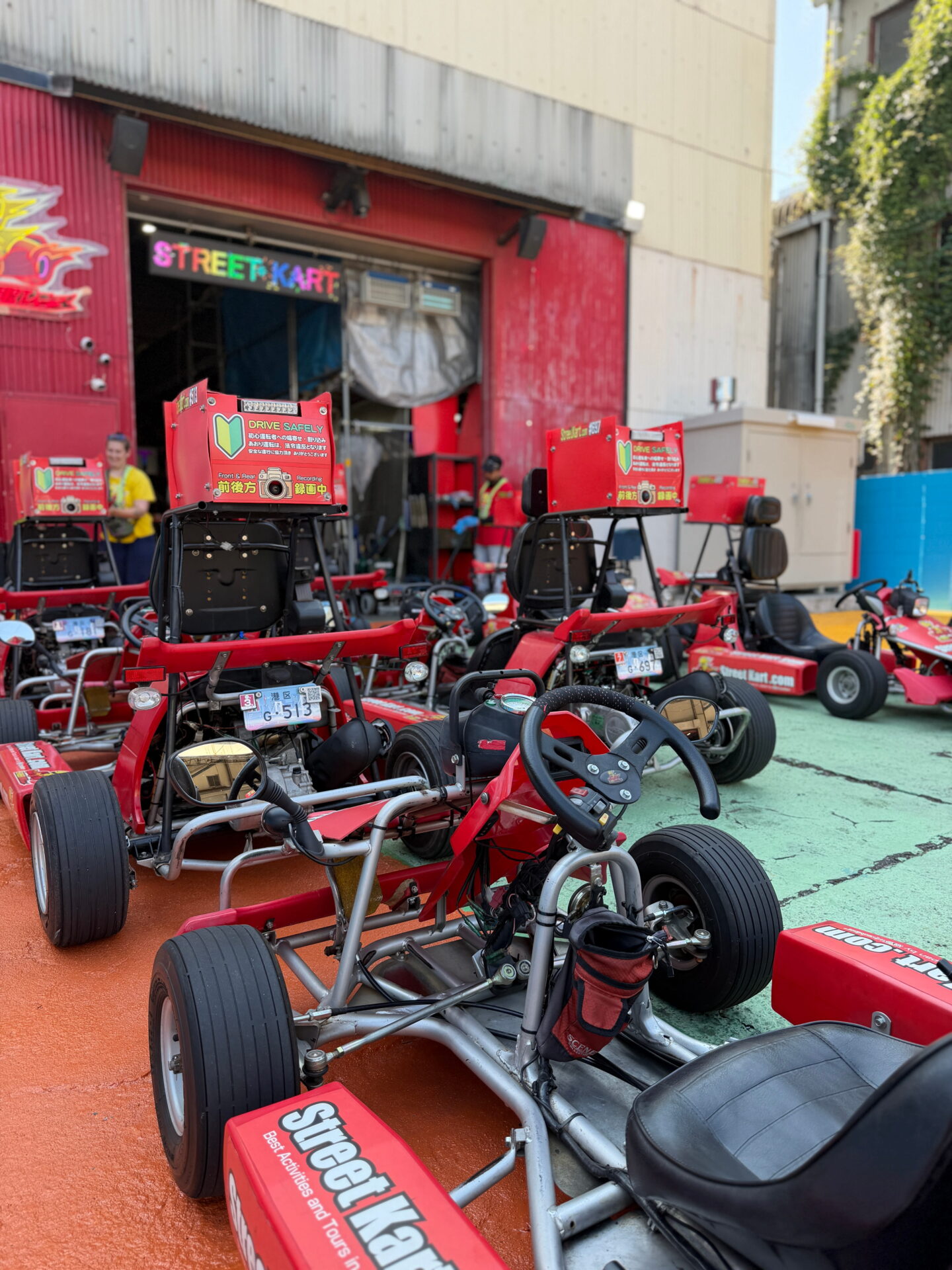 Rows of red Street Karts parked outside the Street Kart garage in Tokyo before the group ride. The open frame karts have low seats steering wheels mirrors and signs reminding drivers to drive safely.