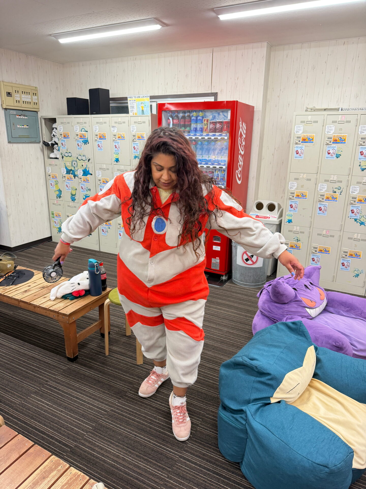 Nishi tries on a red and grey character onesie inside the Street Kart changing area. Large purple and blue character beanbags sit beside her with lockers and a Coca Cola vending machine behind her.