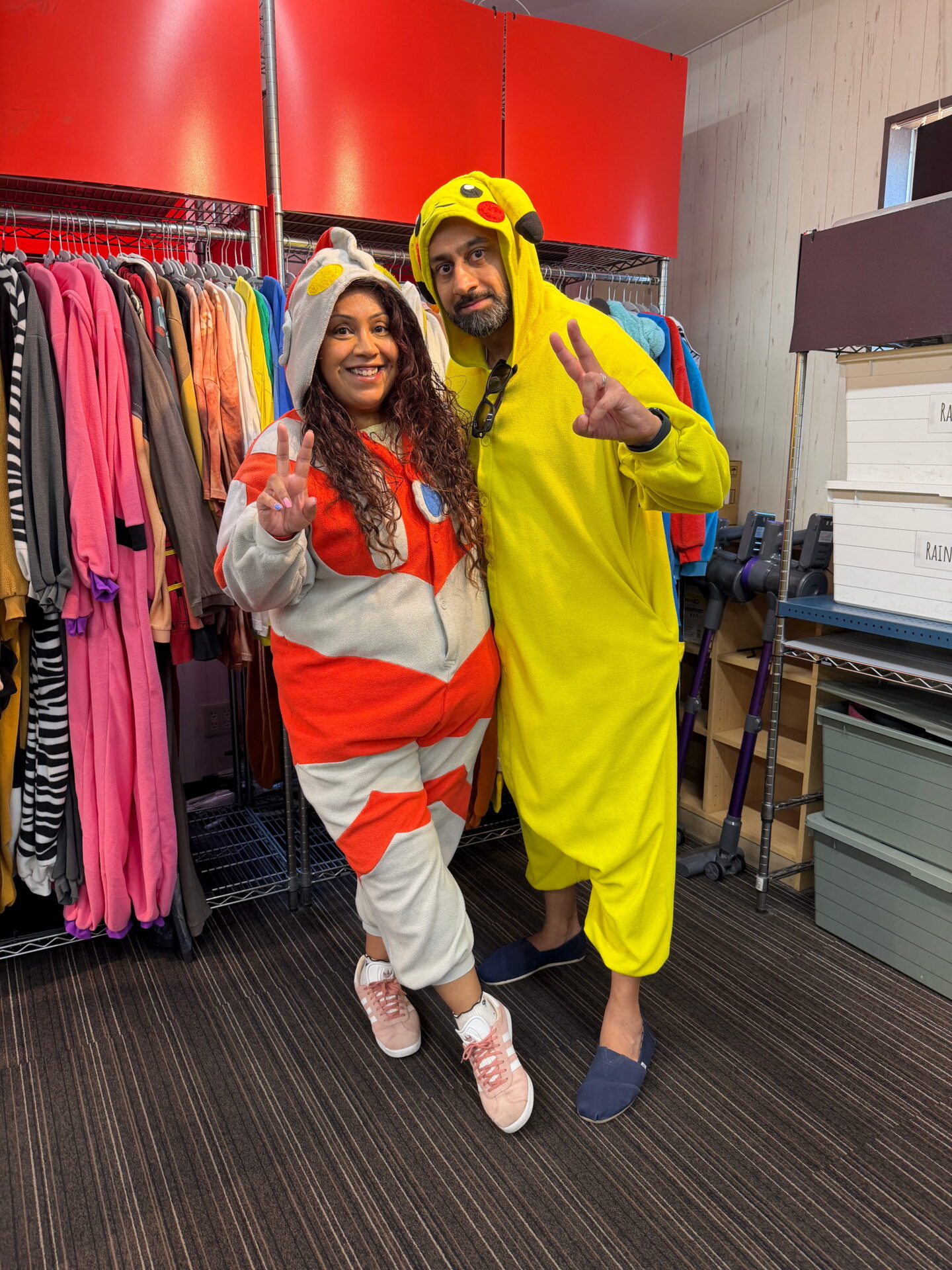 Nishi and Jass pose together in front of racks of colorful costumes before their Street Kart ride. Nishi wears a red and grey character onesie while Jass wears a bright yellow Pikachu costume and both hold up peace signs.