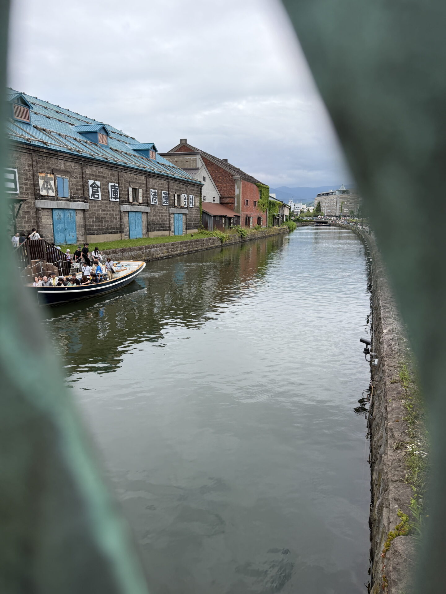 A sightseeing boat carrying tourists travels along Otaru Canal in Hokkaido. Historic stone warehouses with bright blue doors and roofs line the water beneath a cloudy sky with mountains visible in the distance.