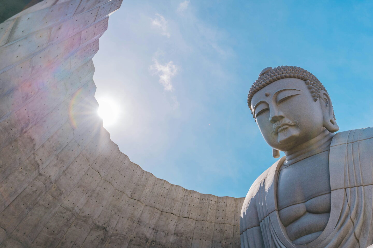 A towering stone Buddha statue at the Hill of the Buddha in Sapporo, photographed from below against a bright blue sky. Sunlight streams over the curved concrete walls surrounding the statue.