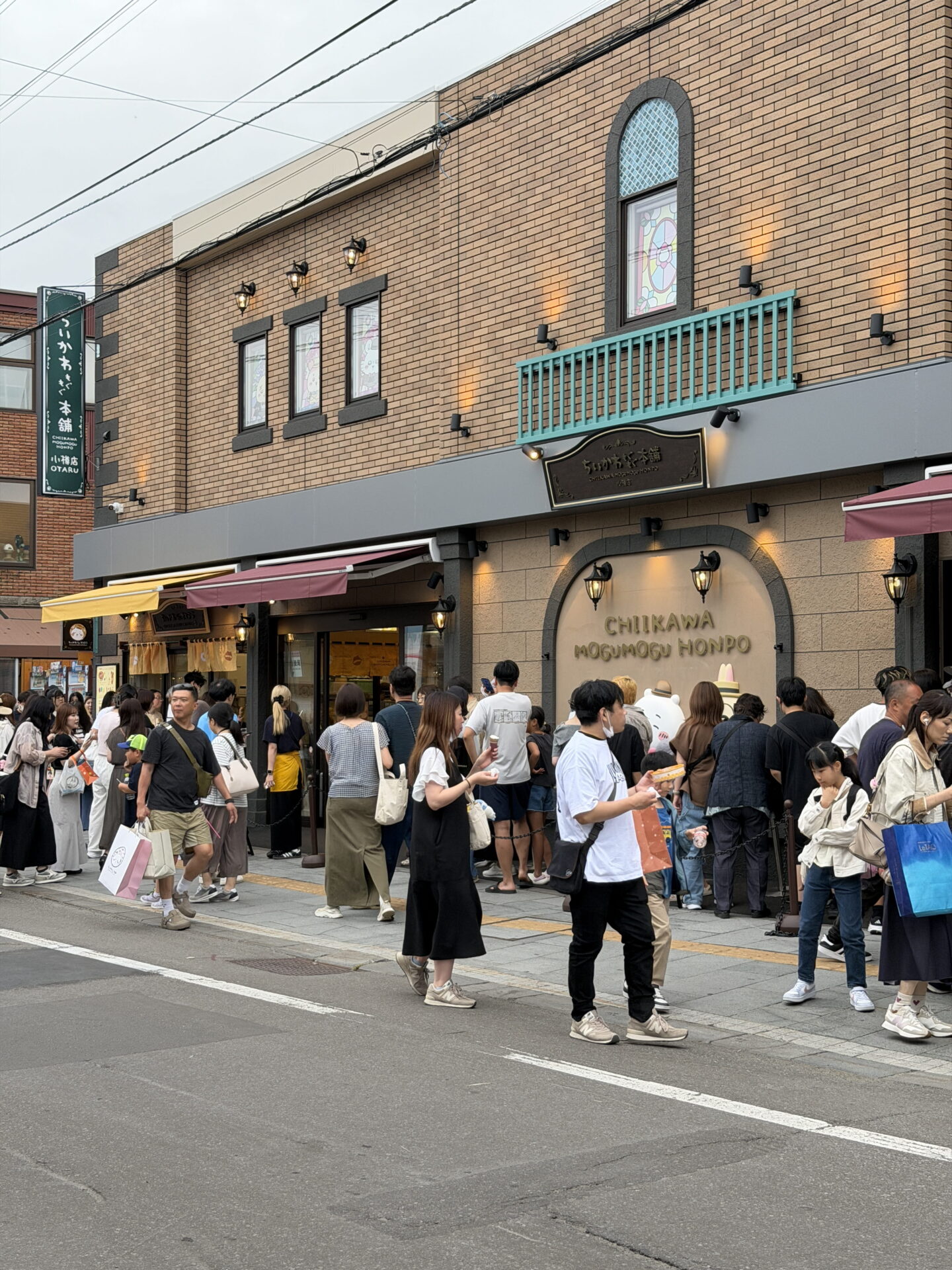 Crowds of shoppers gather outside Chiikawa Mogumogu Honpo in Otaru on a busy shopping street. The two storey brick building features a large sign reading "CHIIKAWA MOGUMOGU HONPO" above the entrance and a green vertical sign with Japanese lettering.