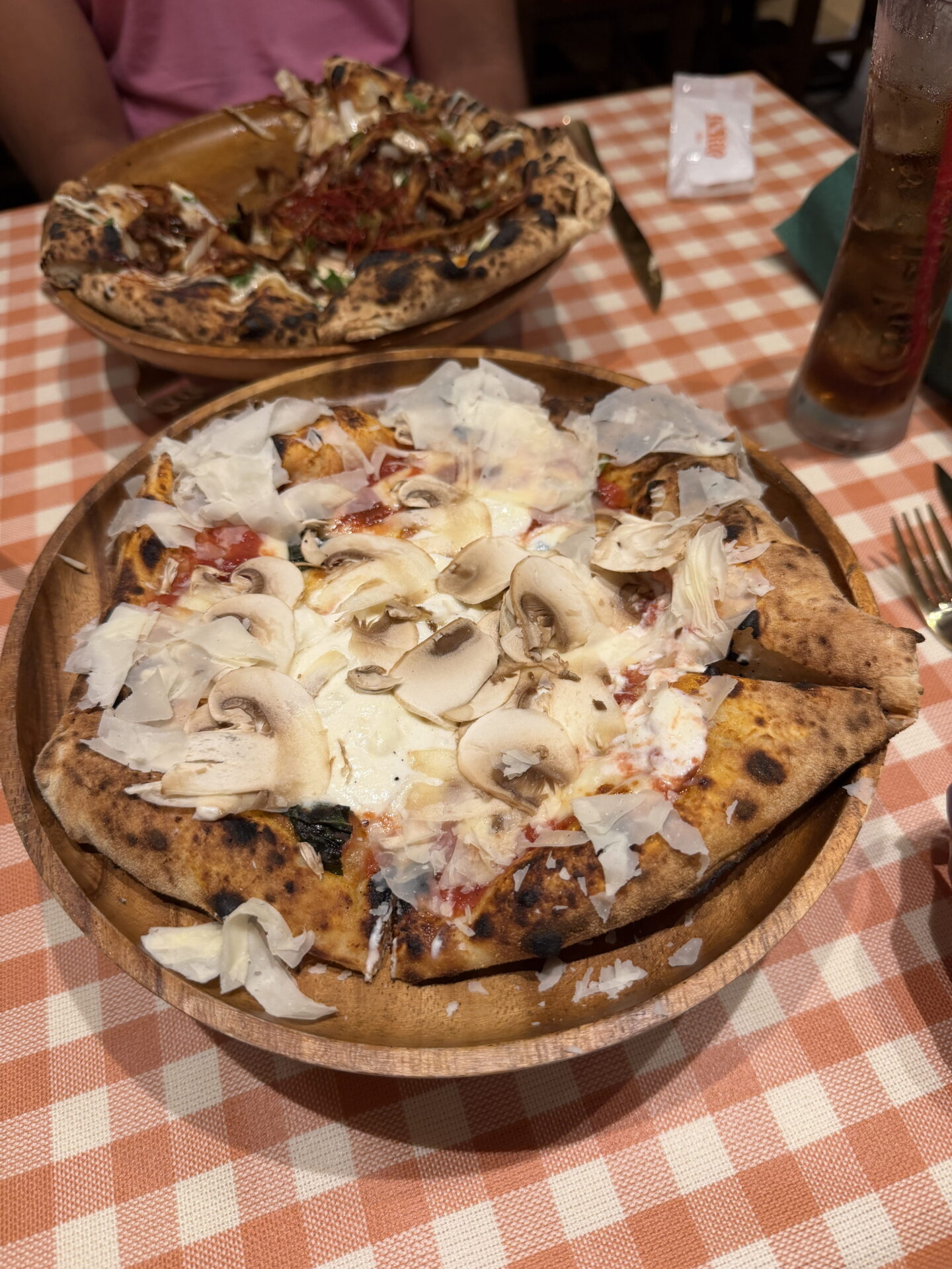 Wood fired pizza topped with sliced mushrooms, melted cheese, tomato sauce and generous shavings of Parmesan on a round wooden serving board. A second pizza sits behind it on an orange and white chequered tablecloth.