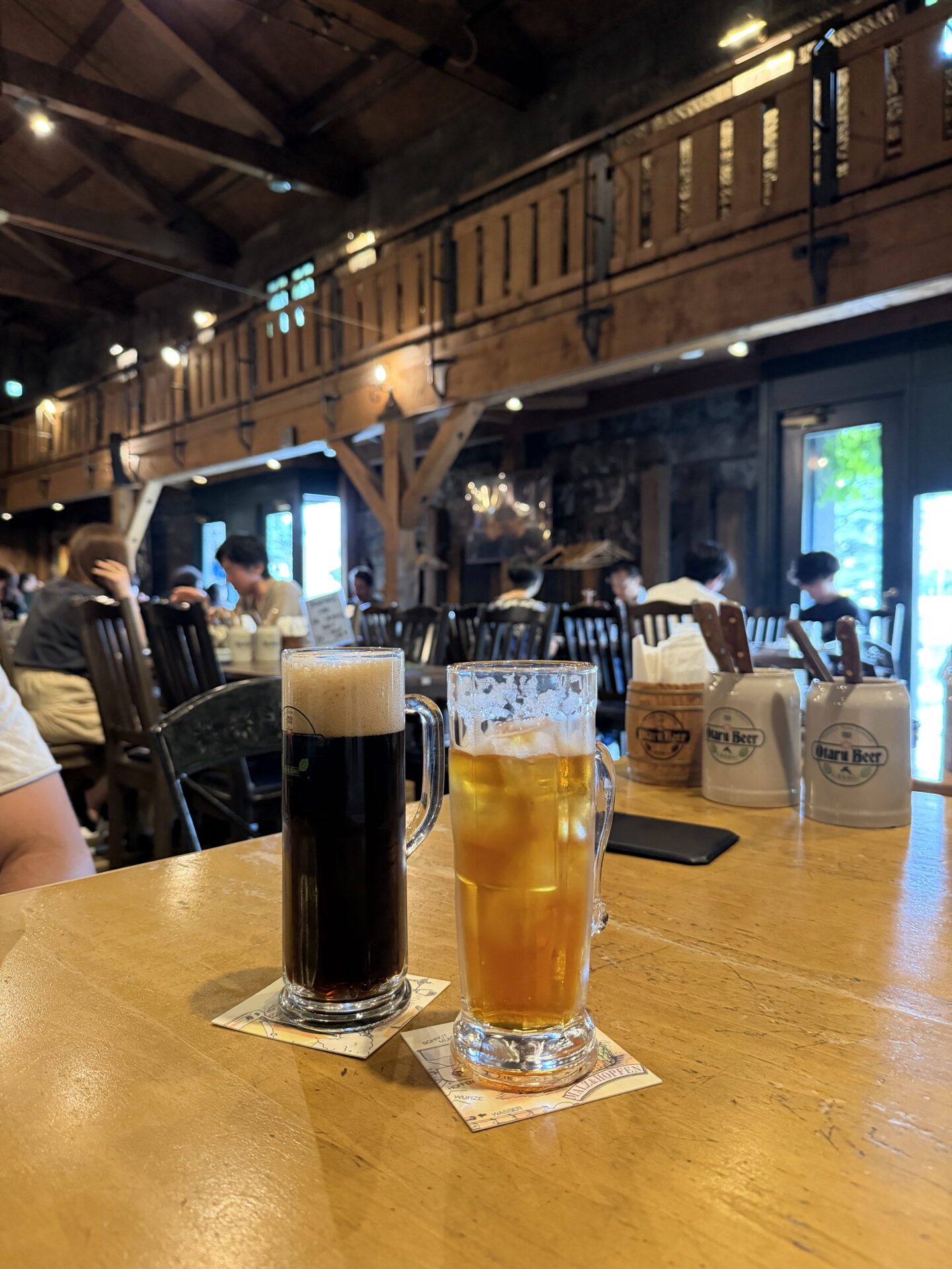 Two large glass beer mugs sit on a wooden table inside a rustic beer hall in Otaru. One contains dark beer with a thick foamy head while the other holds a golden drink with ice. Diners sit at wooden tables beneath exposed beams and a mezzanine.