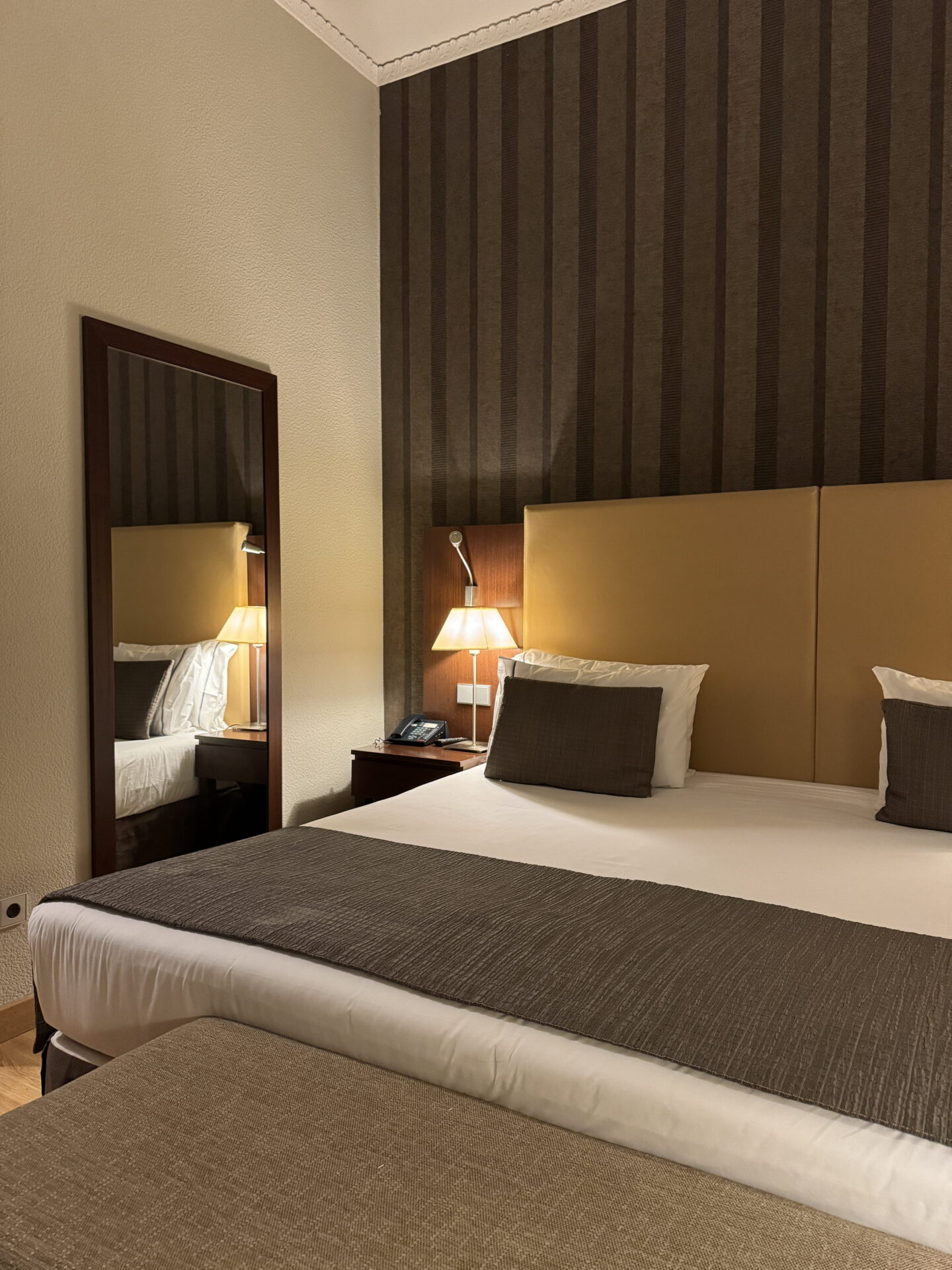A neatly made hotel bed with white bedding, dark brown cushions and a matching bed runner against a beige upholstered headboard and striped brown wallpaper. A bedside lamp glows beside a wooden framed mirror and a cushioned bench sits at the foot of the bed.