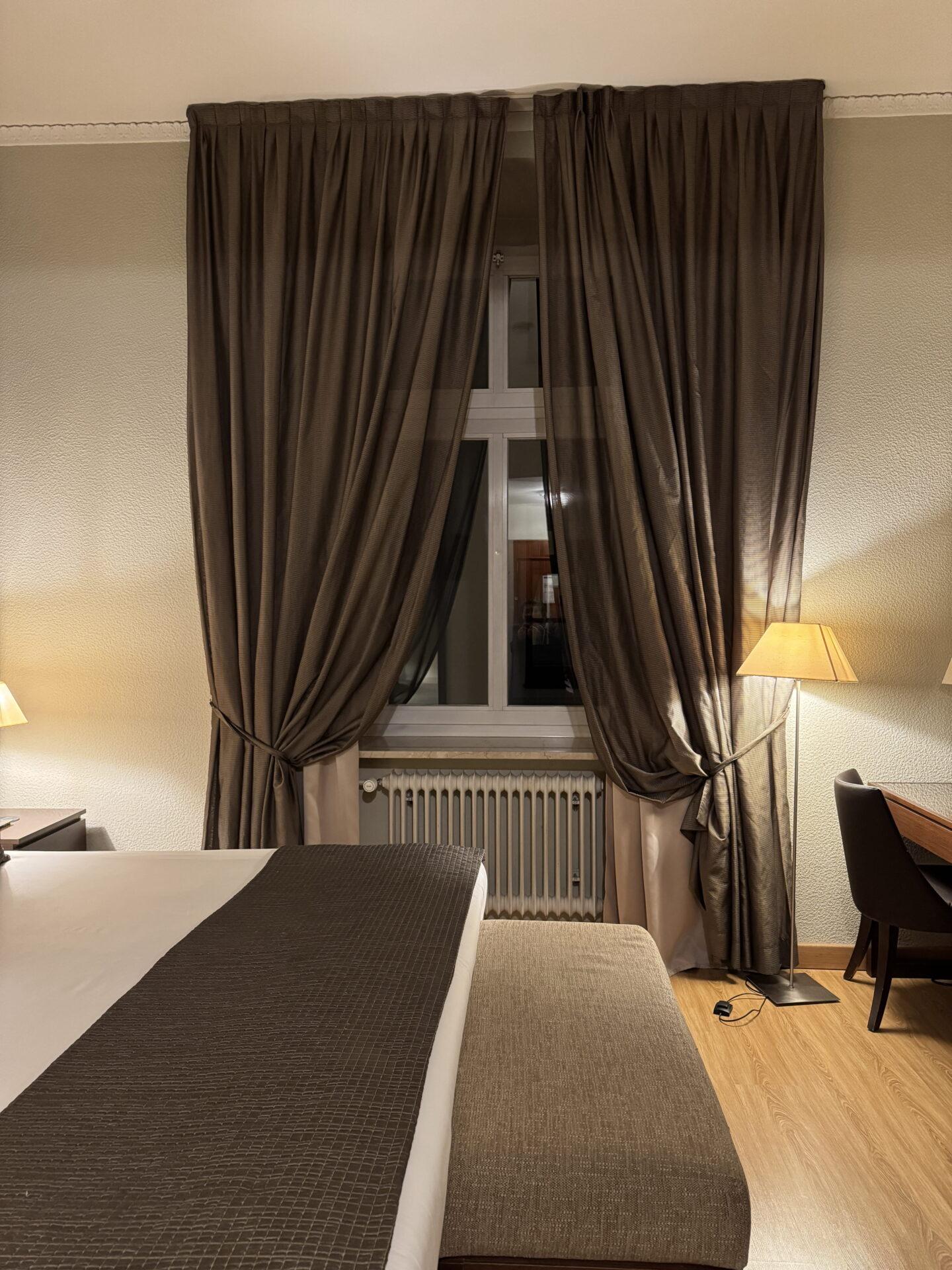 A hotel bedroom with a white bed, dark brown runner and upholstered bench facing a tall window framed by heavy brown curtains. A floor lamp illuminates a wooden desk and chair beside the window.