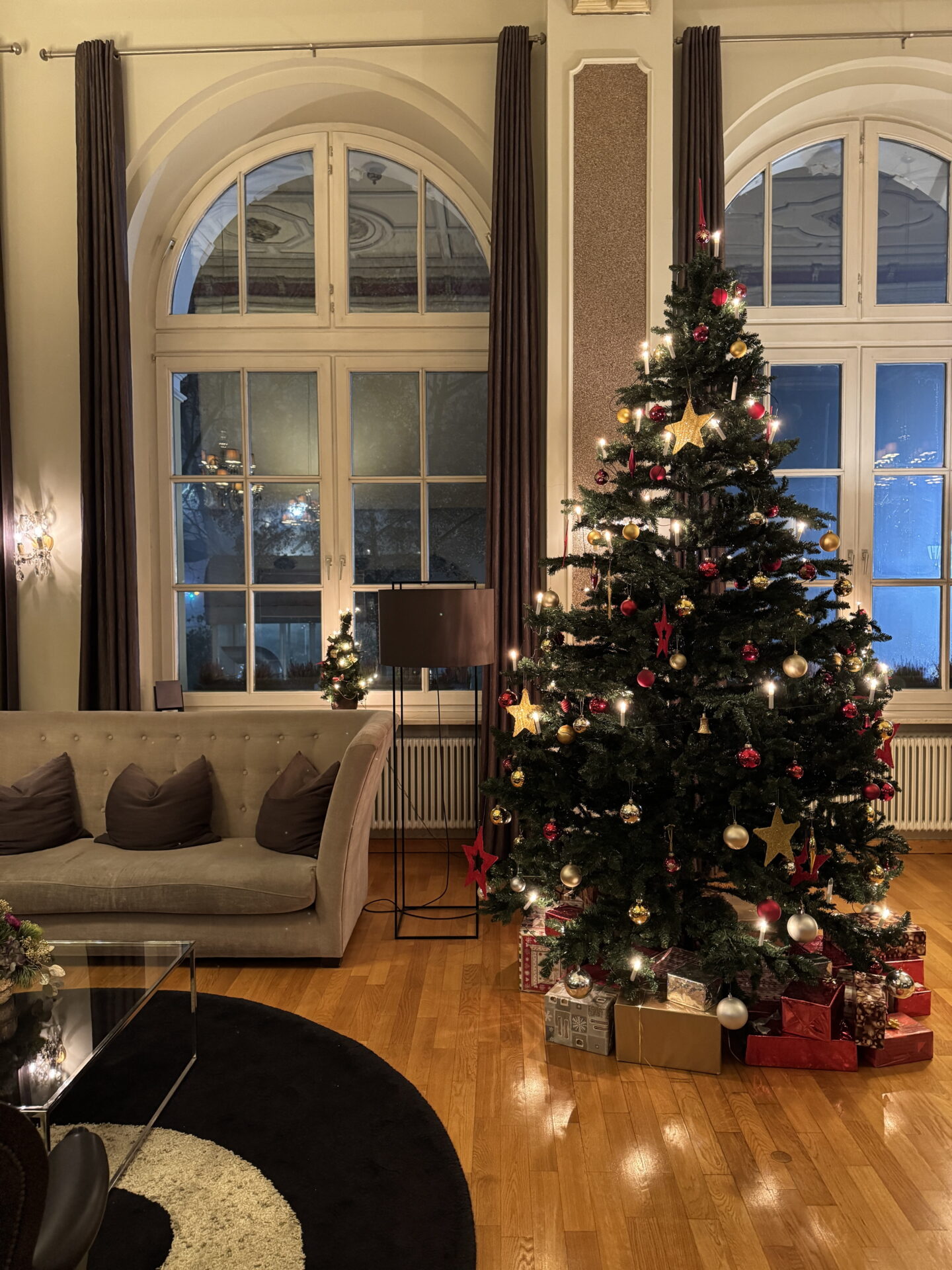 A tall Christmas tree decorated with glowing lights, red and gold baubles and star ornaments stands between two large arched windows in a hotel lounge. Wrapped presents surround the base of the tree beside a beige sofa and glass coffee table.