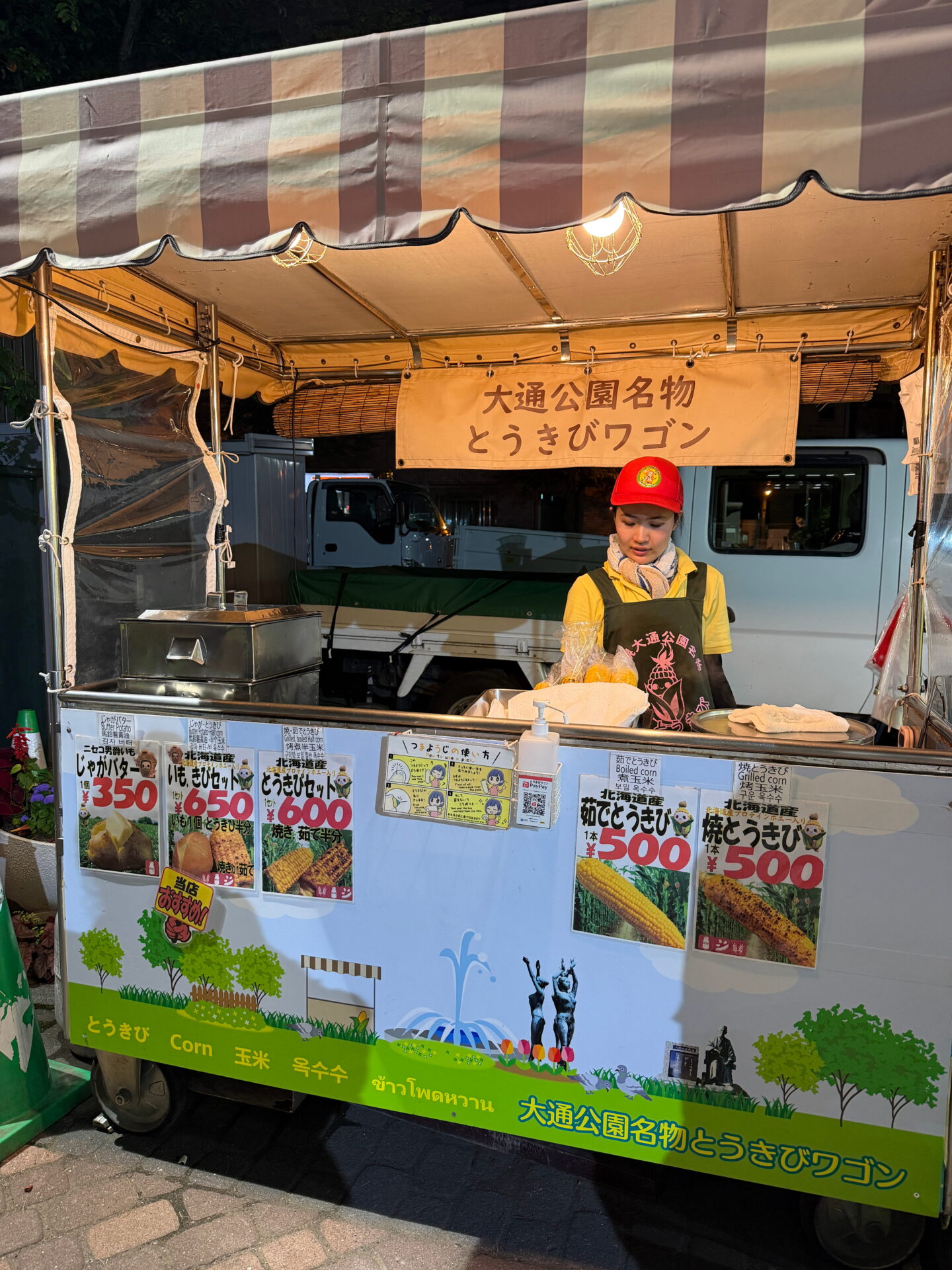 A street food vendor selling Hokkaido sweetcorn at a stall in Odori Park in Sapporo. Signs advertise boiled corn and grilled corn for 500 yen each, alongside baked potatoes and corn sets.