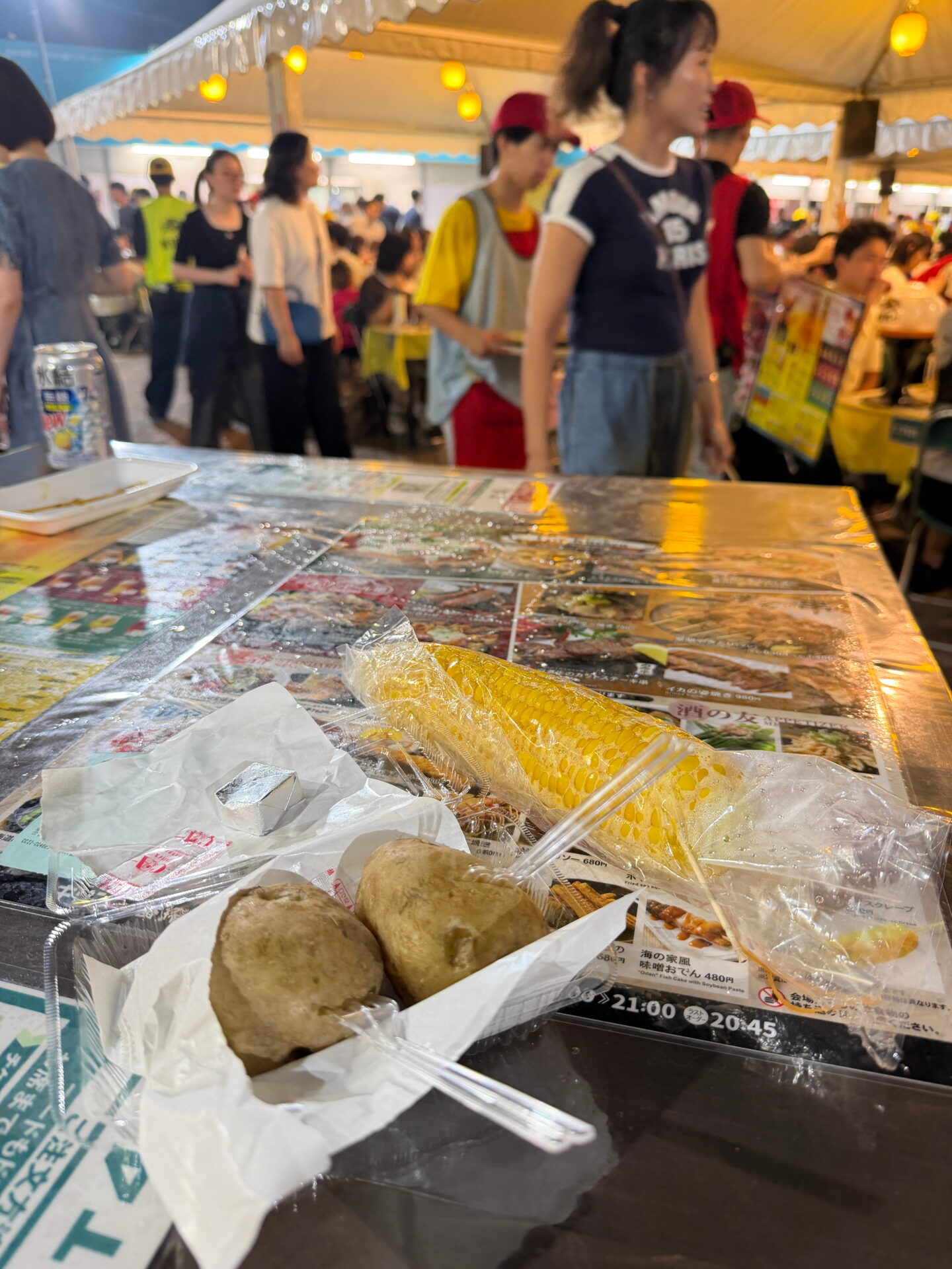 Two baked potatoes and a yellow corn on the cob served in takeaway packaging on a table at a busy food tent in Sapporo. Diners and food vendors fill the background beneath warm yellow hanging lights.