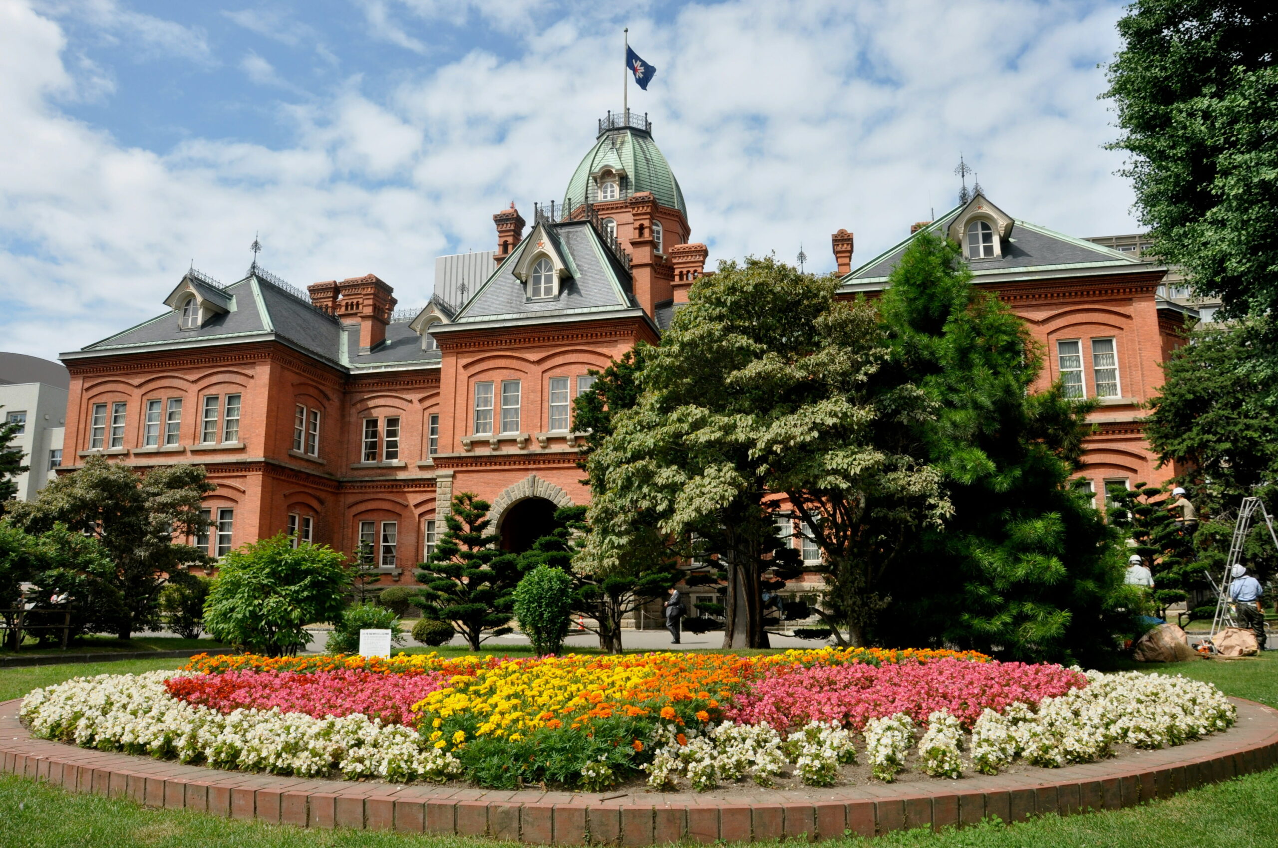 The historic red brick Hokkaido Government Office in Sapporo with its distinctive green domed roof and Japanese flag flying above. A colourful flowerbed filled with pink, yellow, orange and white flowers stretches across the foreground.